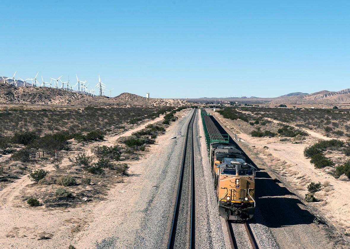A long freight train approaches between a wind-turbine farm and 