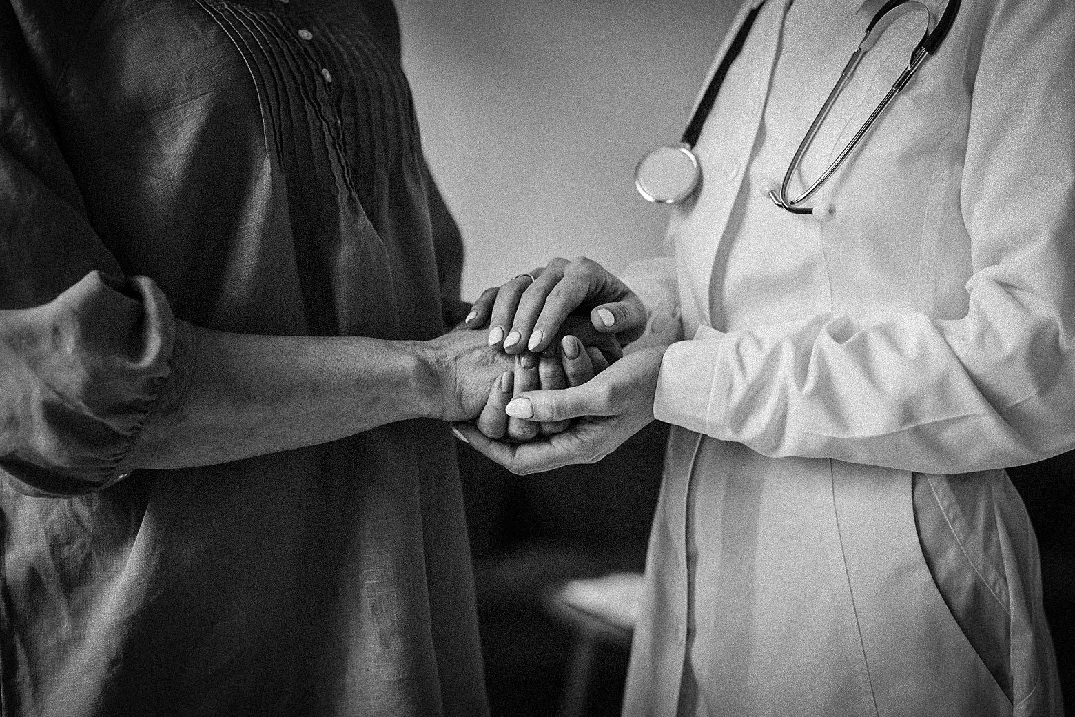 Black-and-white image of a doctor in a white coat and stethoscope holding the hand of a civilian, dressed in a dark top.