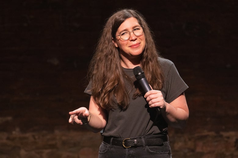A woman in a black t-shirt and glasses stands on a stage with a microphone, pointing at the audience.