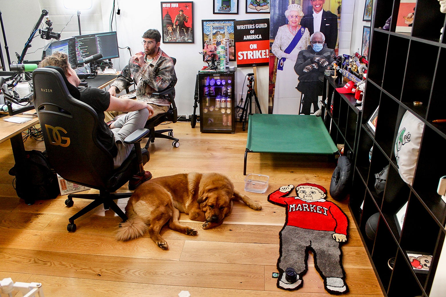 Two men have a conversation in front of several computer monitors, with a dog lying on the ground beside them.