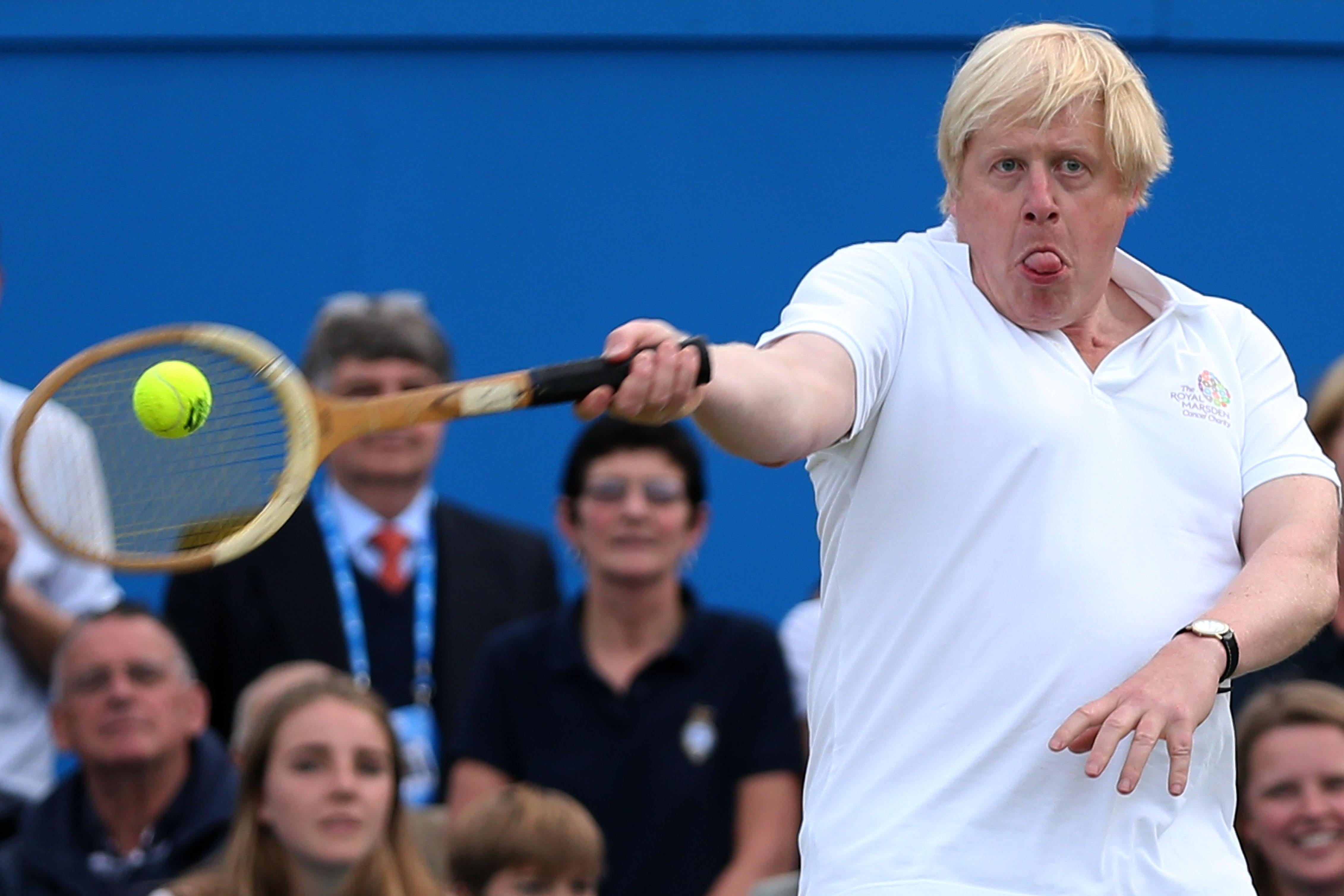 Boris Johnson sticks his tongue out as he hits a tennis ball while spectators watch.