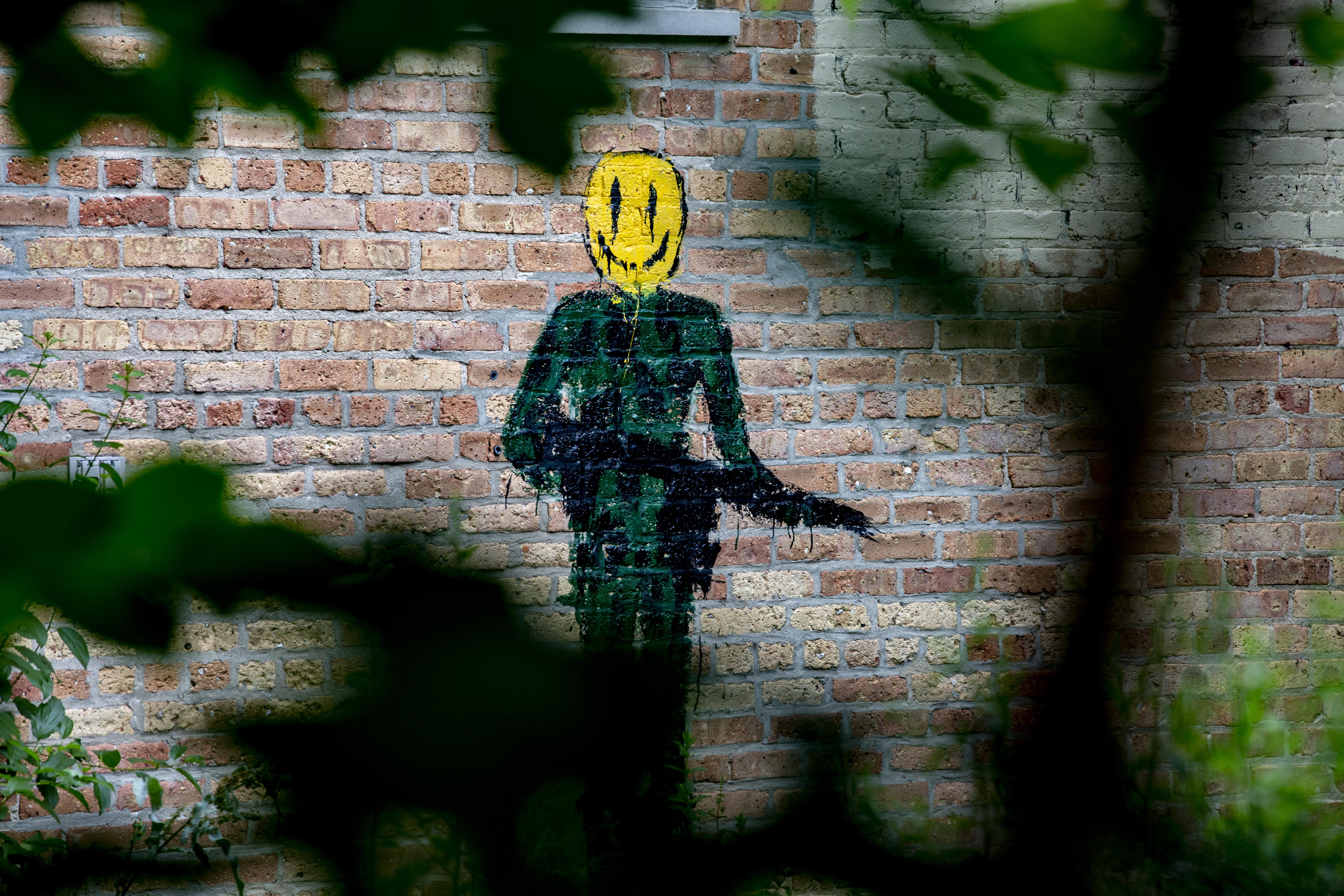 Through foliage, we see brick wall with a mural of a person in fatigues, with a yellow smiley face for a head, holding an automatic weapon.