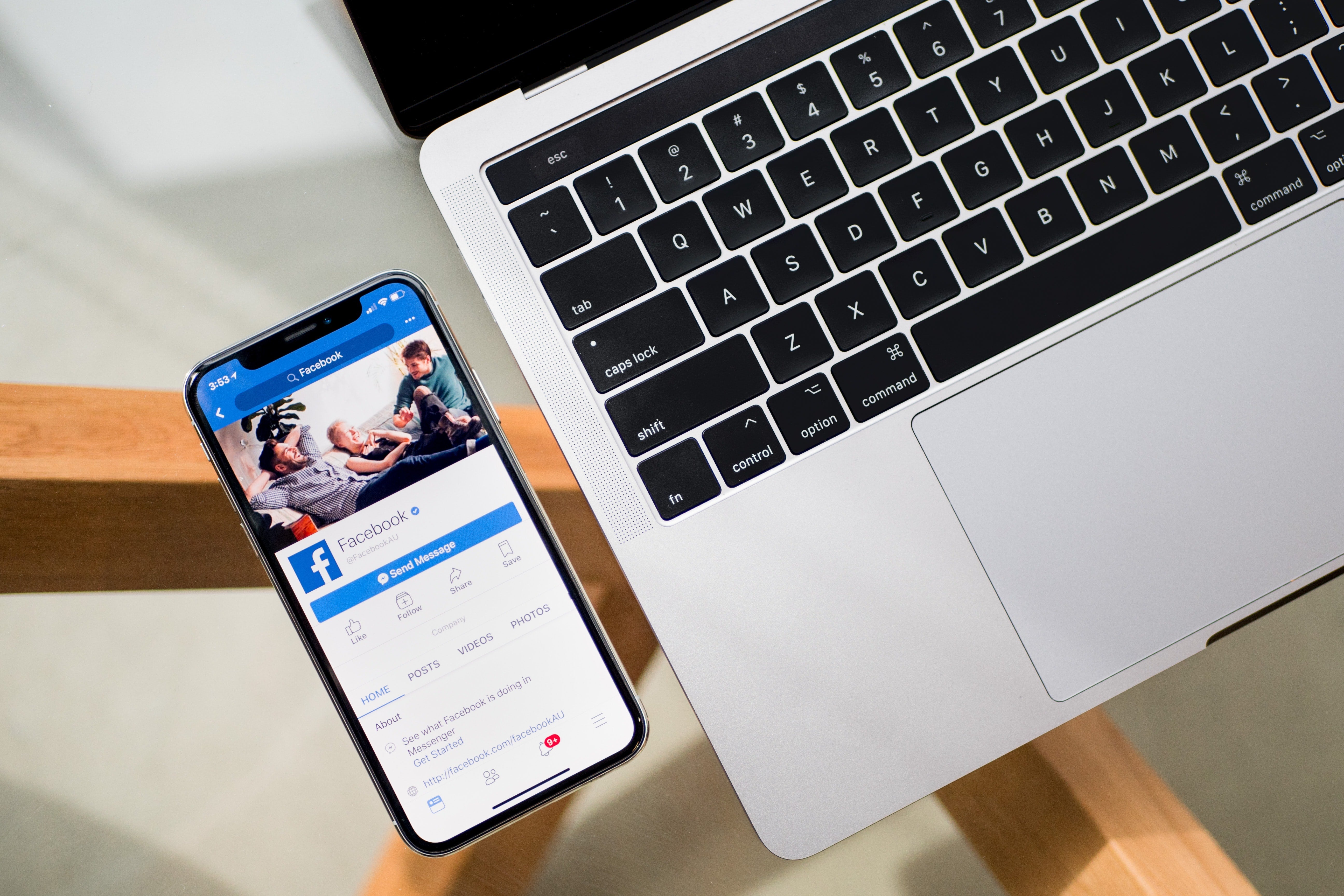 A phone displaying Facebook sits on a glass table next to a laptop.