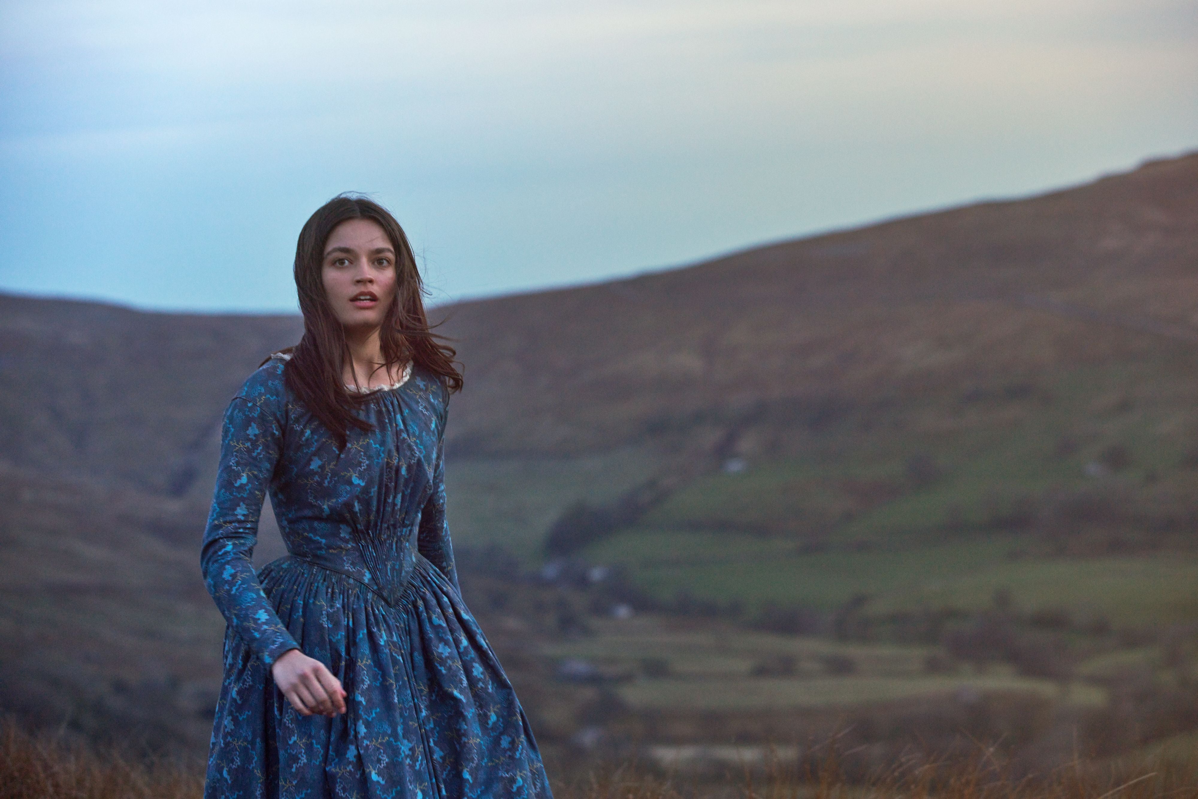 A slim attractive white woman with her long brown hair parted down the middle stands in a blue-green floral dress on a hill above the English countryside, the wind in her hair and her lips just barely parted