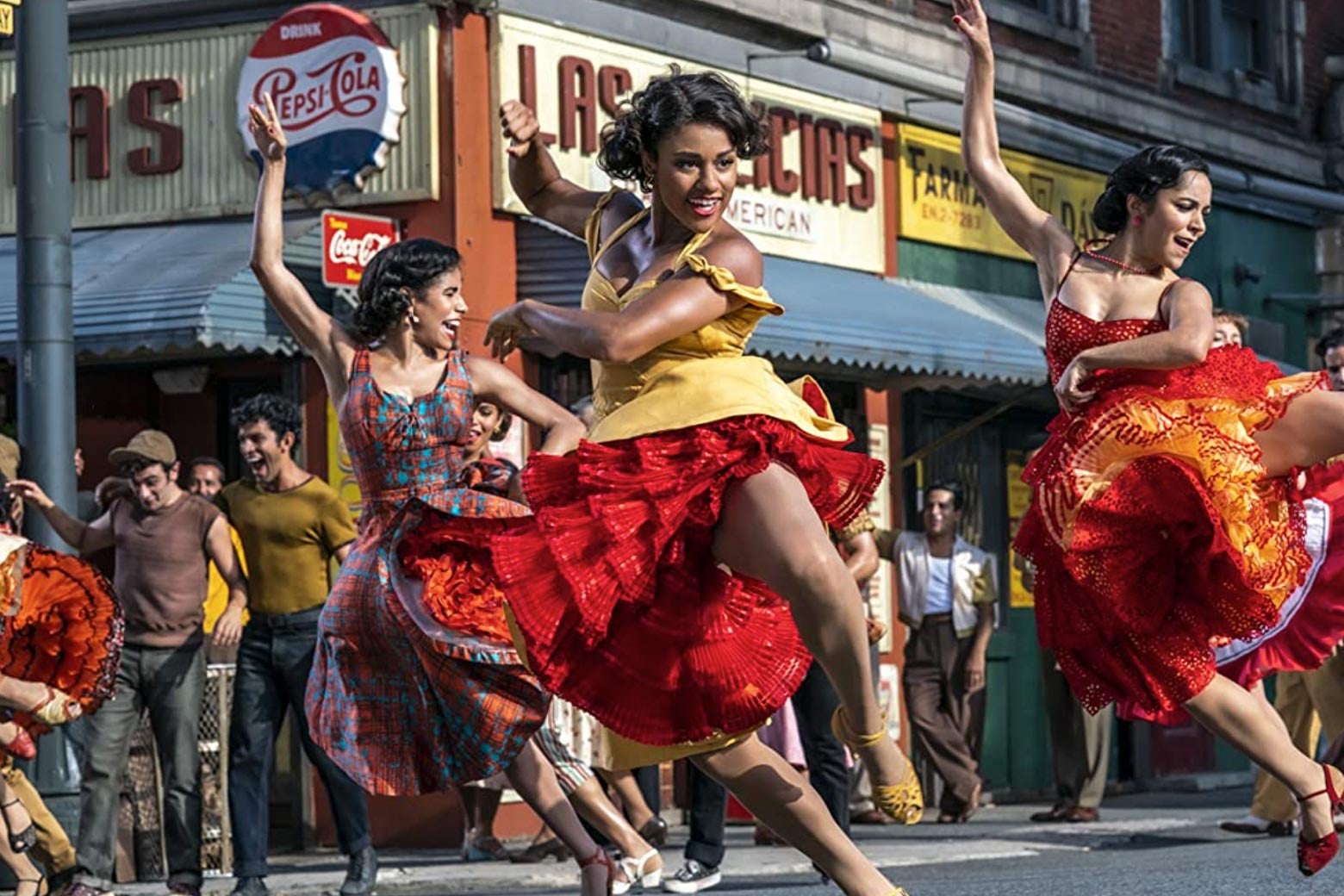A still from the 2021 West Side Story of three women in colorful dresses dancing in a 1960s-era New York street