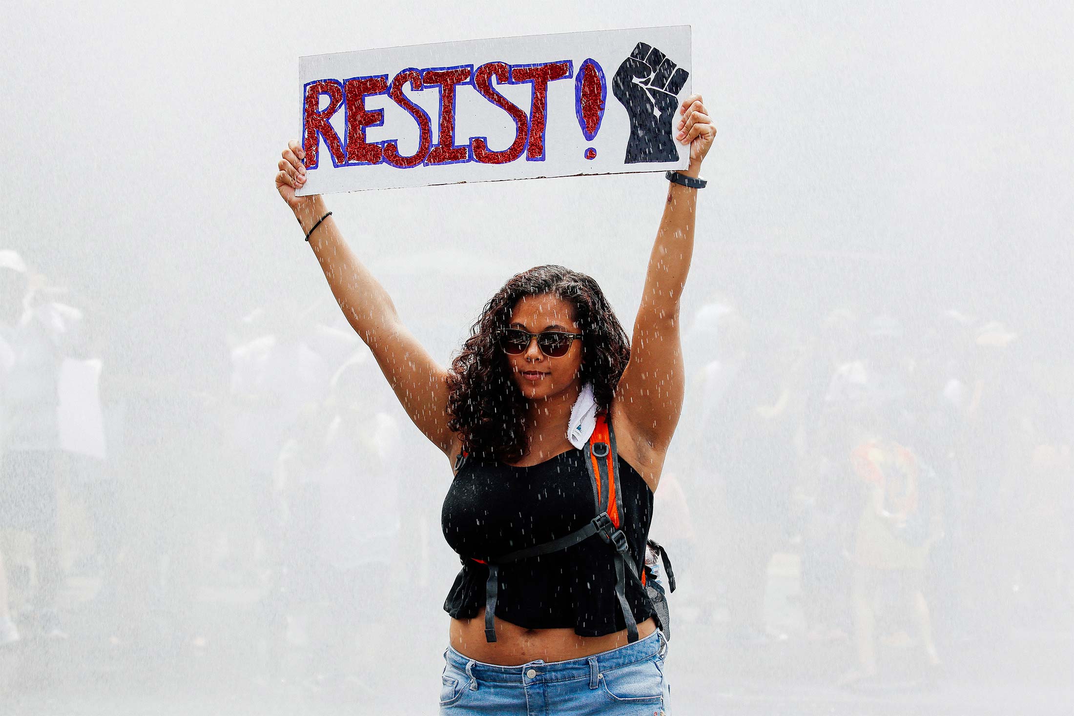 A young woman defiantly holds up a "RESIST!" sign as she stands in a spray of water from a fire truck during a protest.
