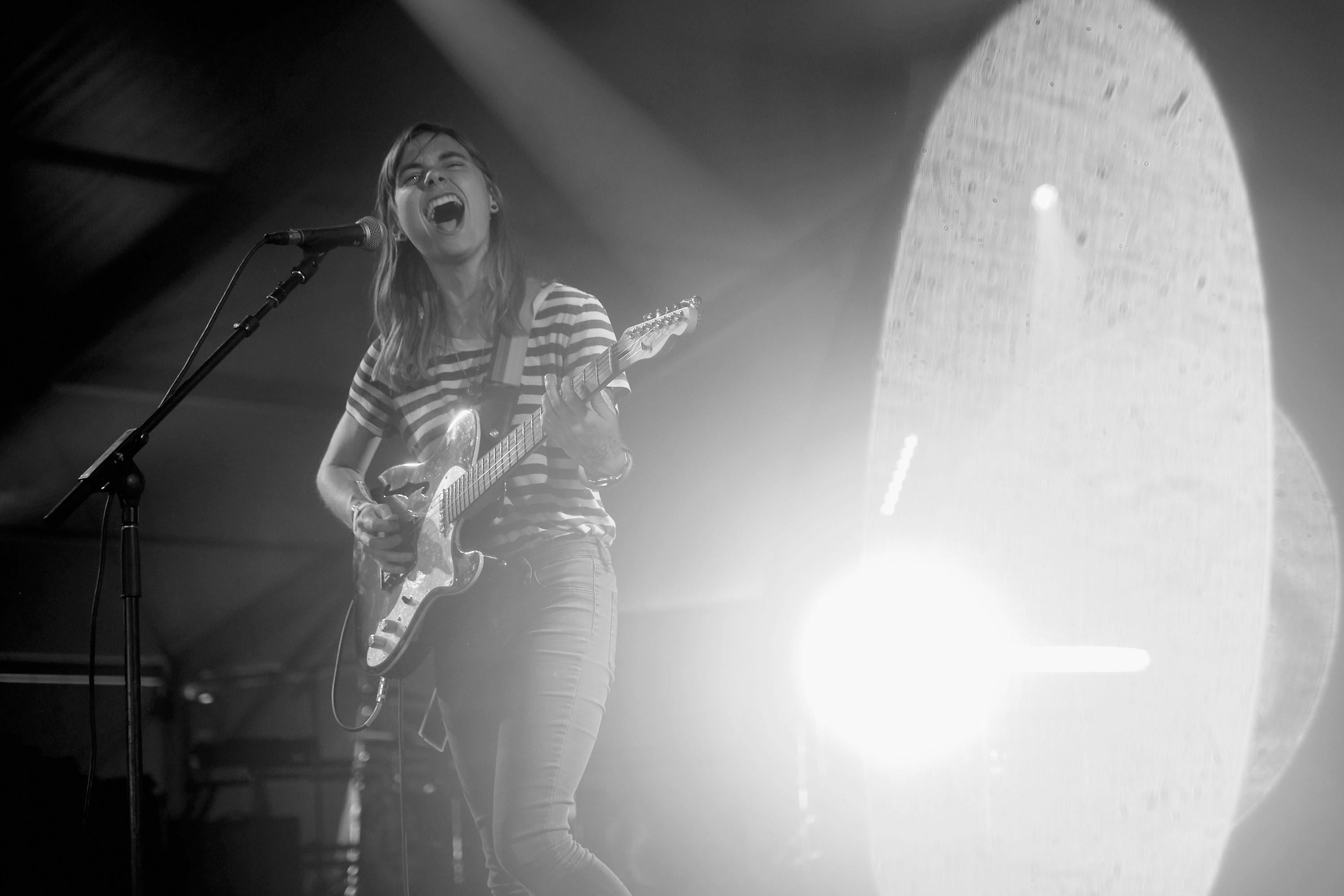 A woman with shoulder-length hair performs on stage. She is playing a guitar and singing while wearing a t-shirt and jeans.