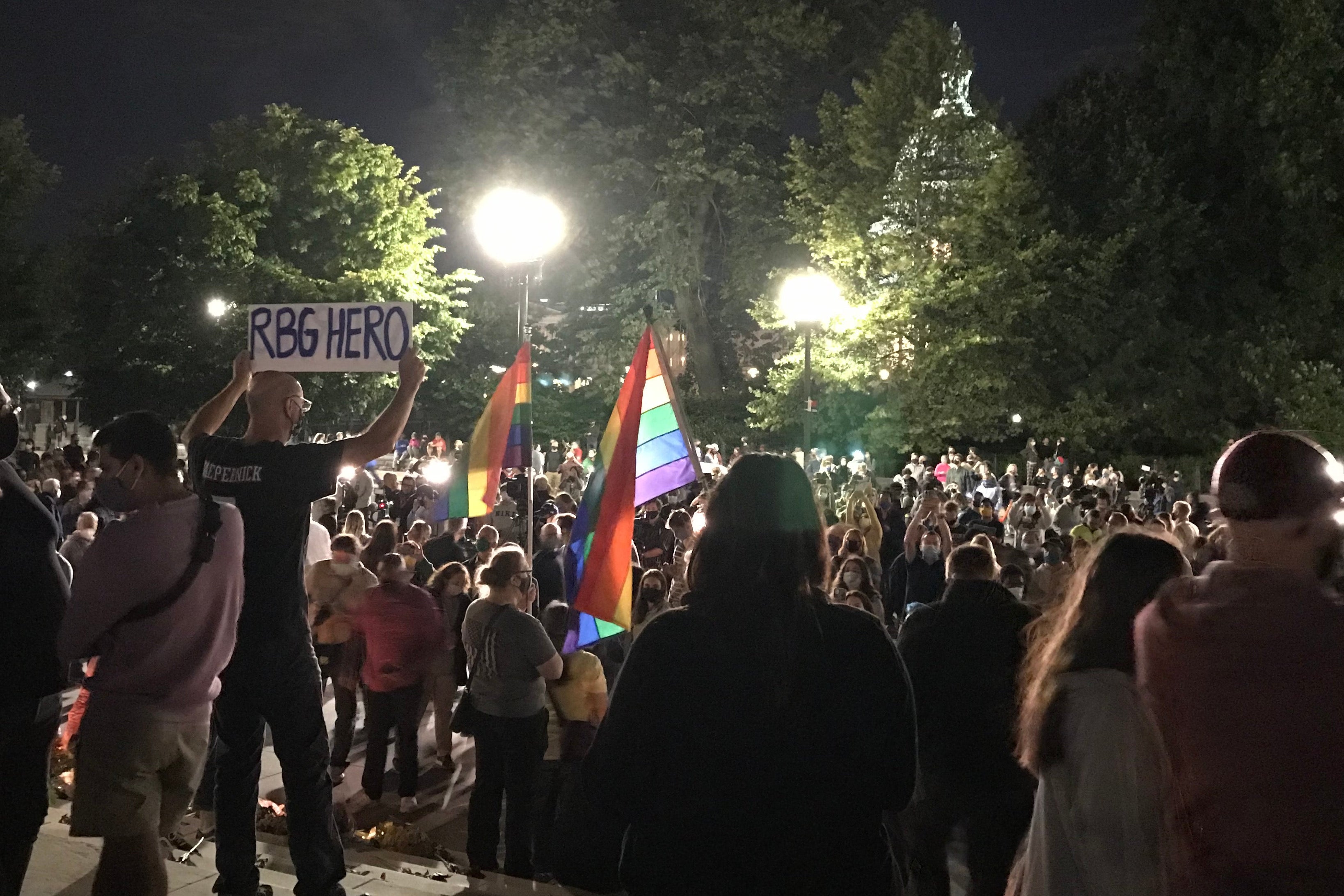 A man holds a sign reading "RBG Hero" amid a large crowd with waving rainbow flags and the Capitol in the background.