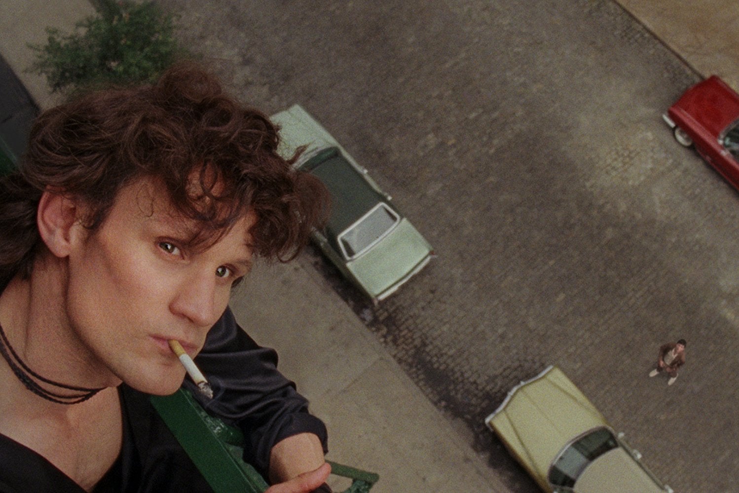 Matt Smith, a young man with curly brown hair and a square jaw, plays Robert Mapplethorpe in the biopic Mapplethorpe. He wears a black silk robe and smoke a cigarette on his fire escape, looking up. Cars move on the street below him.