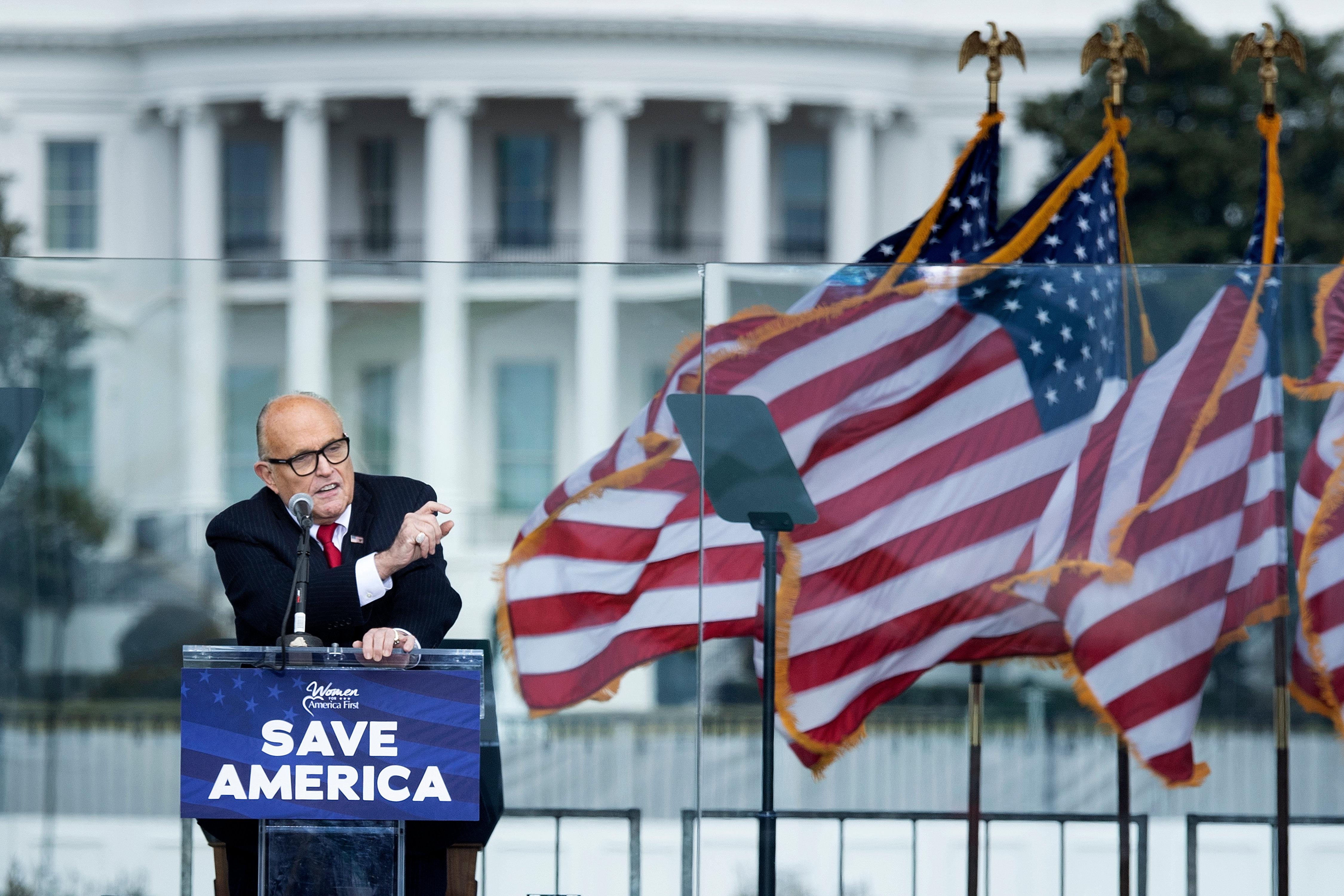 Rudy Giuliani speaks at a podium that says "Save America" with American flags waving beside him. The White House can be seen in the distance behind Giuliani.