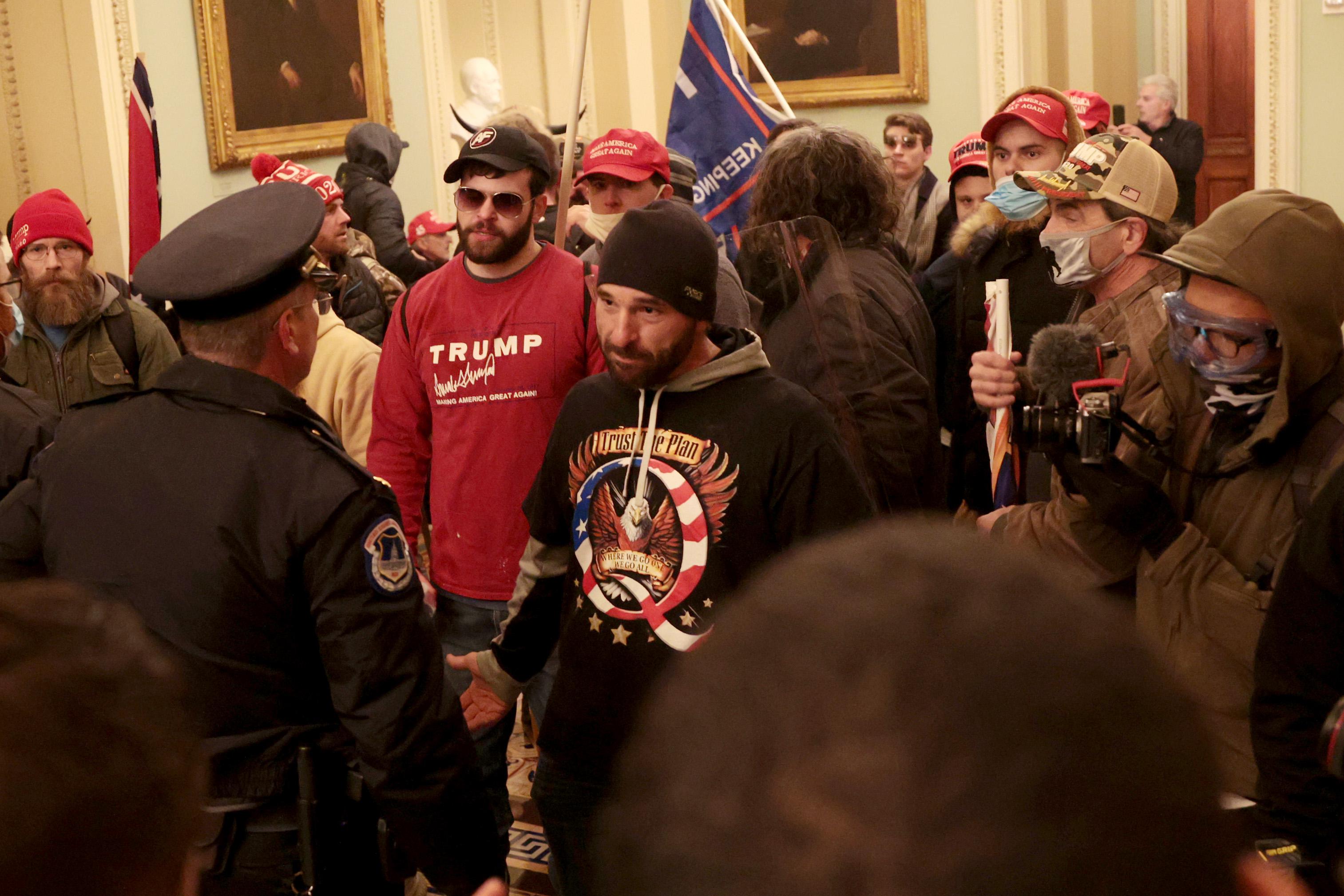 A hallway in the Capitol crowded with rioters wearing Trump insignia. One rioter wearing a black QAnon shirt stares down a police officer.