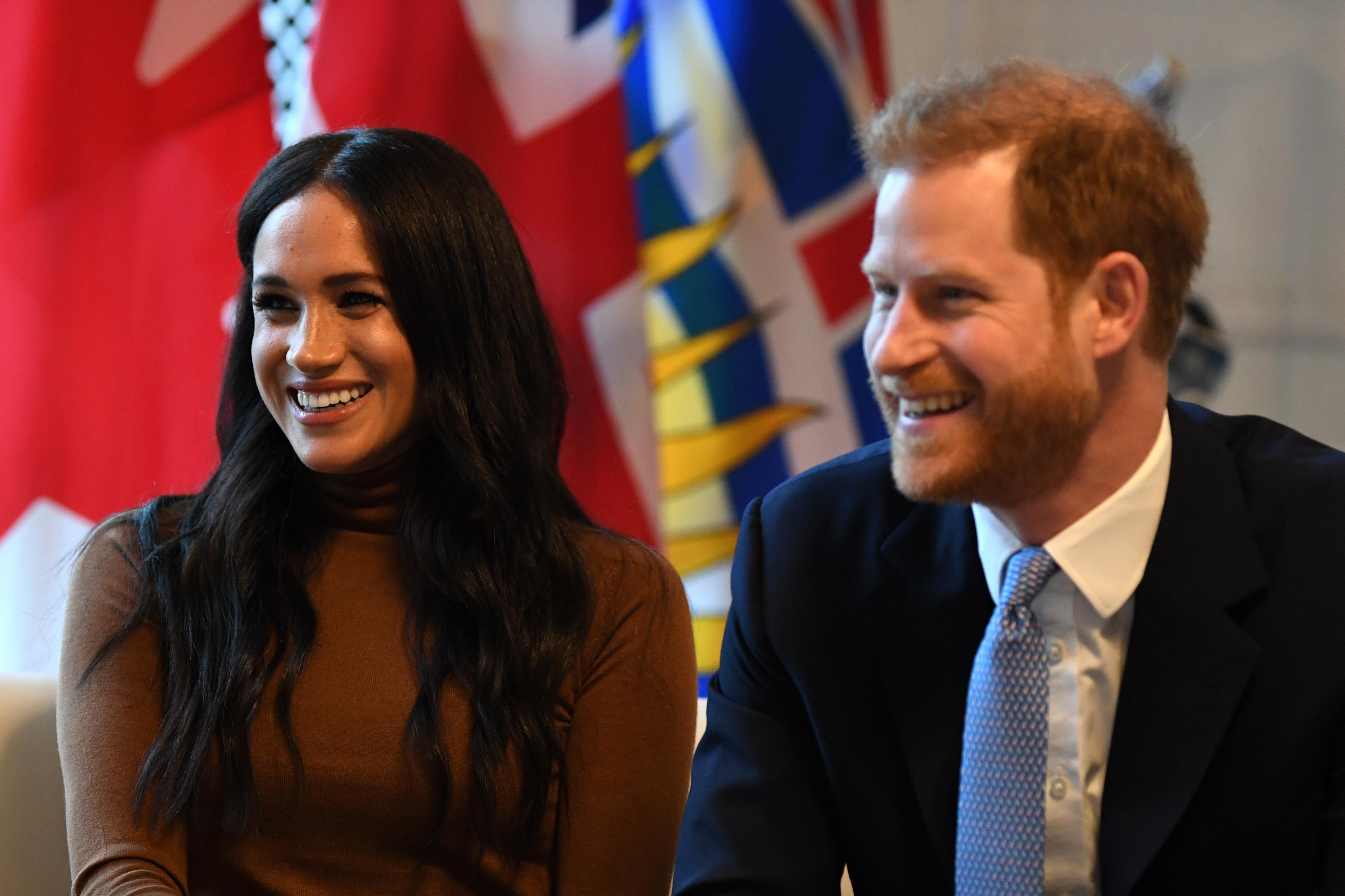 Prince Harry, wearing a navy blue suit, sits next to Meghan Markle, wearing a brown turtleneck. Both of them are smiling.