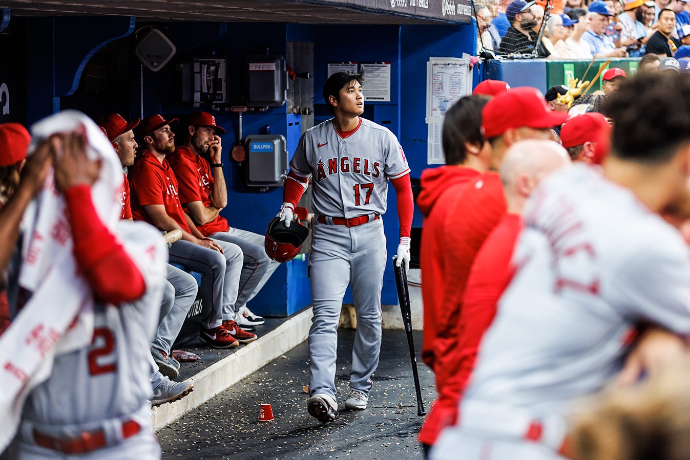 Shohei Ohtani walks in the Angels dugout, looking out at the field, holding his helmet in his right hand and his bat in his left, down to the ground like a cane.