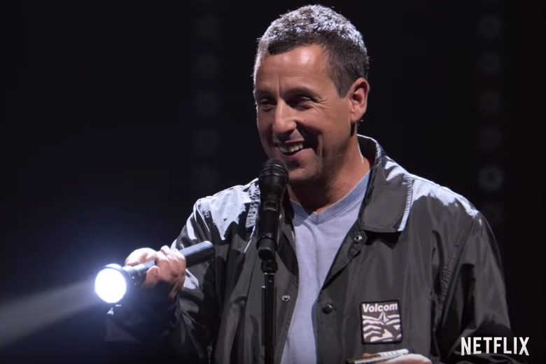 Adam Sandler on stage with a flash light and a microphone.