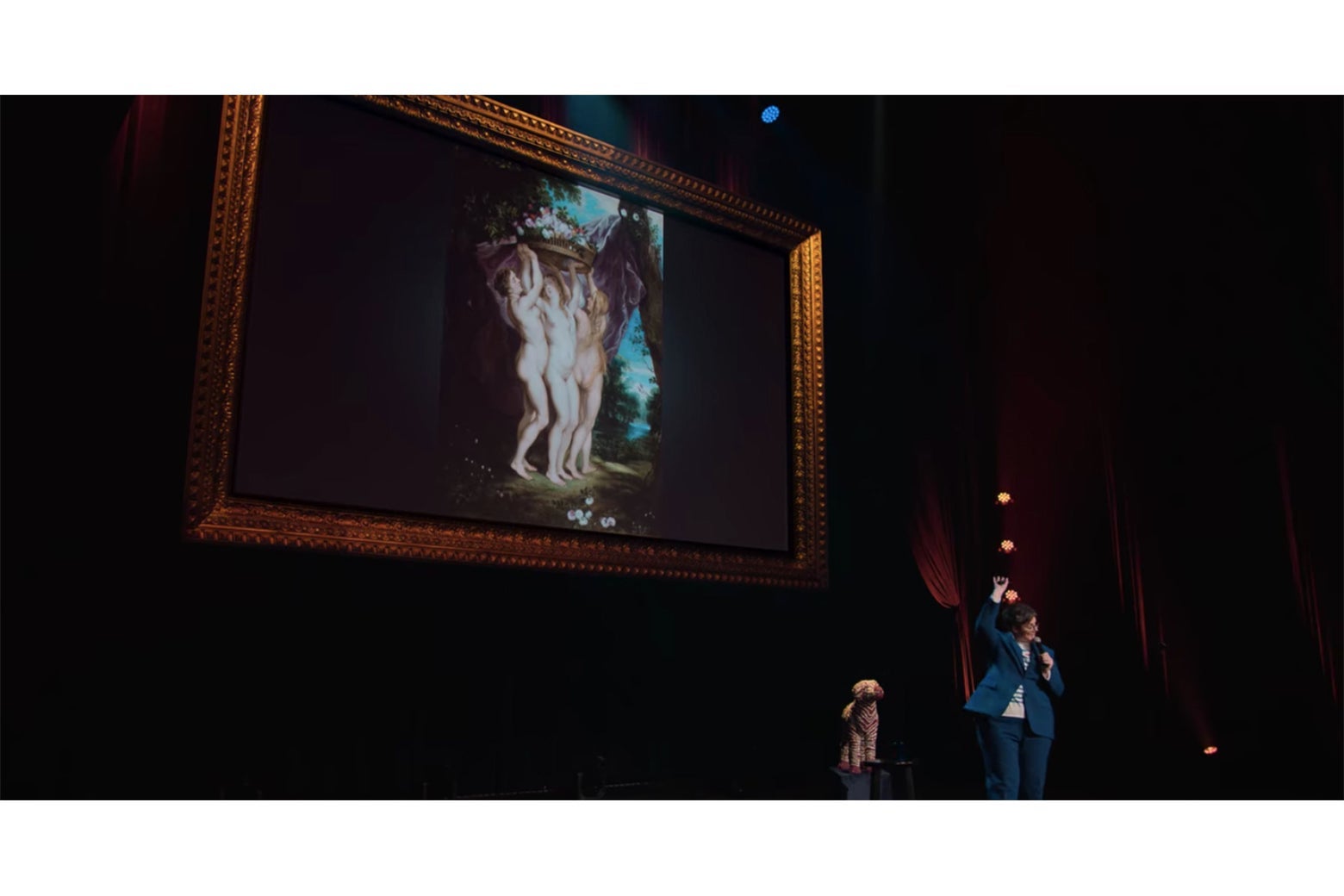 Hannah Gadsby stands in front of yet another painting of three naked women.