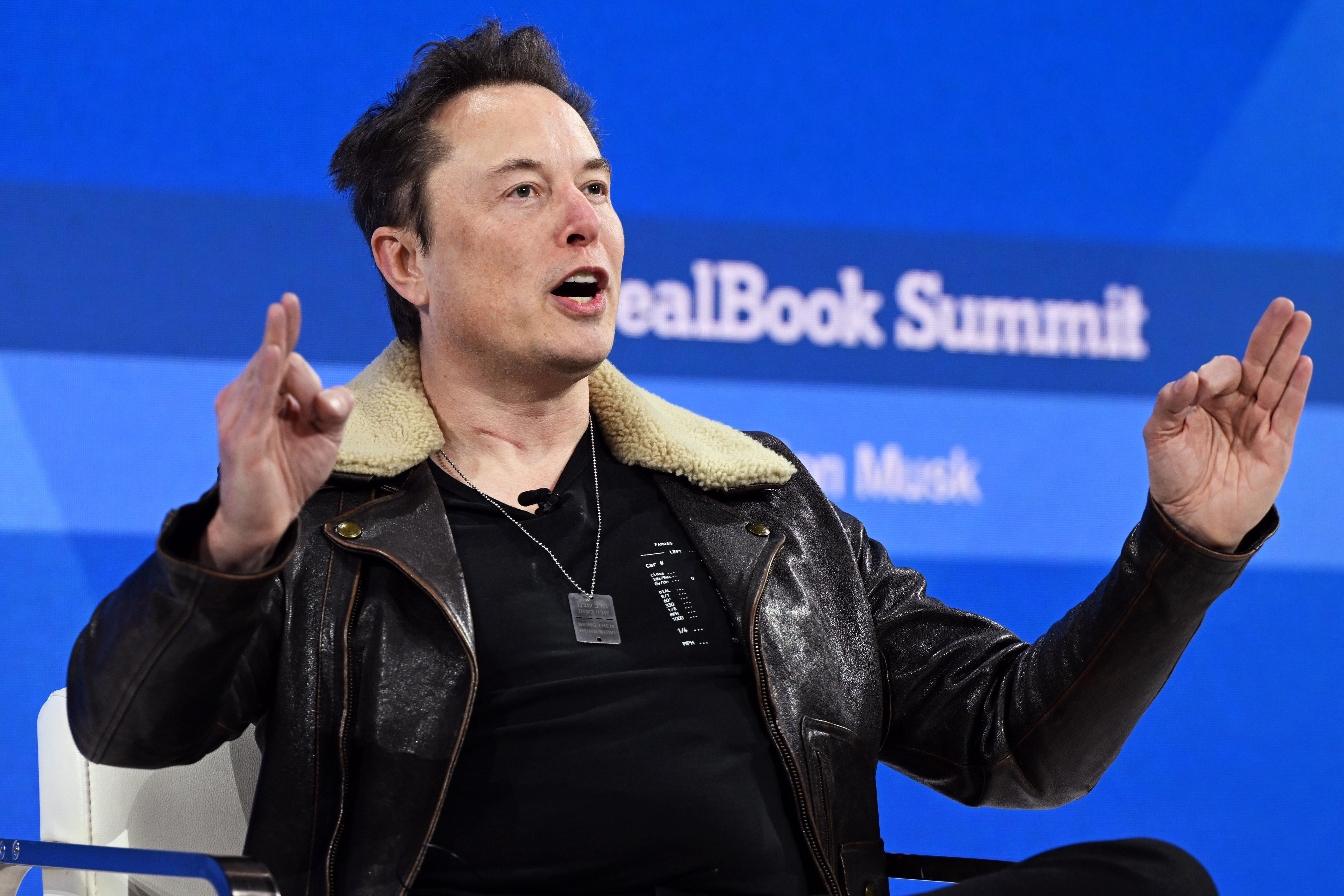 Elon Musk wears a black jacket and speaks animatedly onstage, holding up both hands in an OK symbol.
