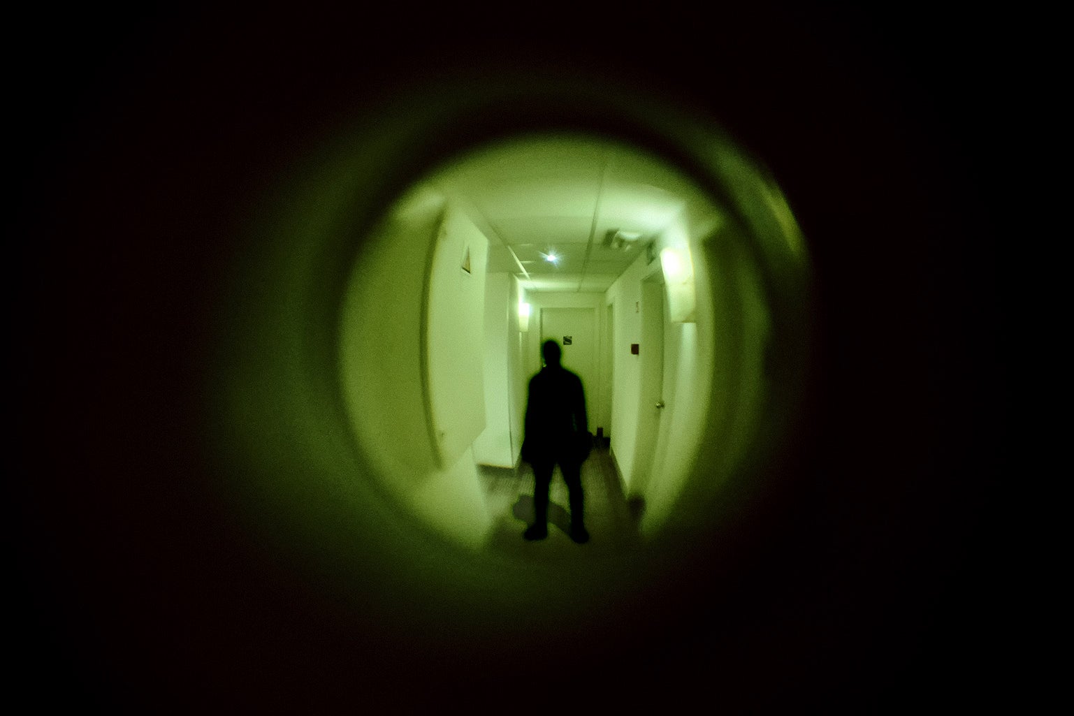 A shadowy silhouetted man is seen through the lens of an apartment door peephole standing in a dimly lit greenish hallway.