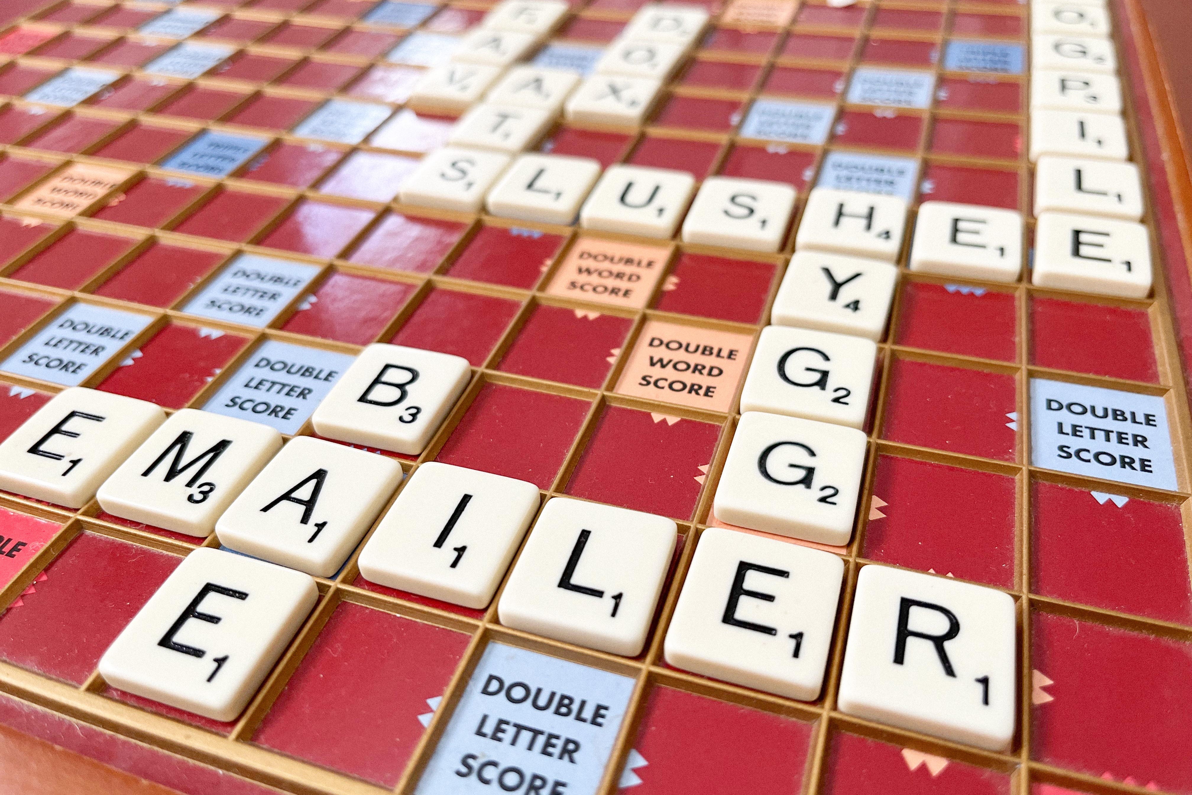 A Scrabble board with some of the new words arranged in a grid on it: Bae, Emailer, Hygge, Slushee, Dogpile, Vax, Ats, Fav, and Dox