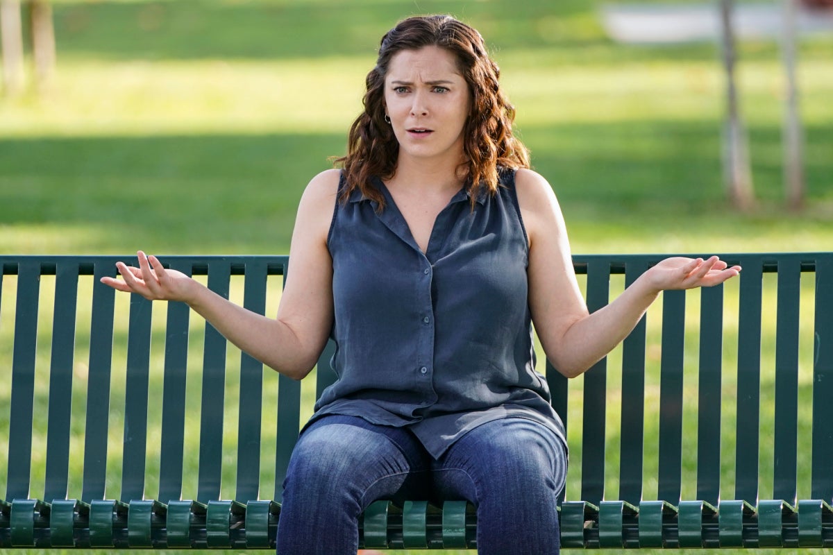 Rachel Bloom sits on a park bench, shrugging her shoulders.