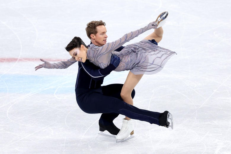Chock skating on her left leg as she bends horizontally forward with her right leg and skate lifted behind her, as Bates crouches holding her with his right leg extended outward parallel to the ice.
