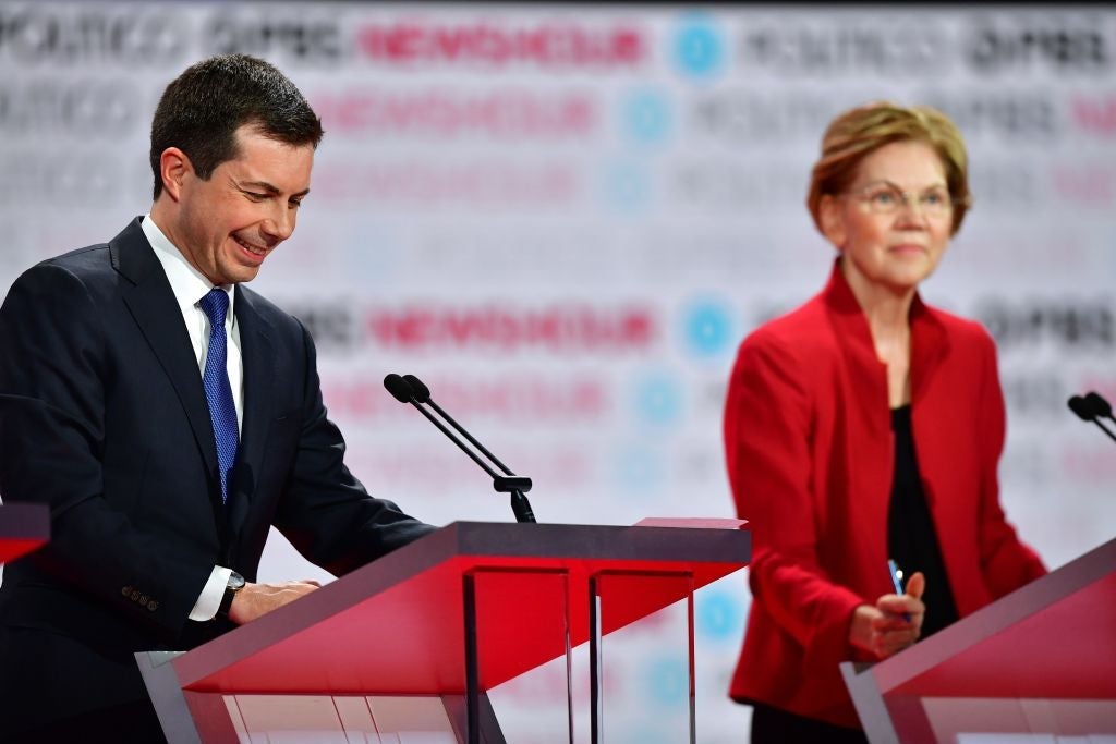 Buttigieg laughs while looking down at his lectern while Warren is seen in the background.