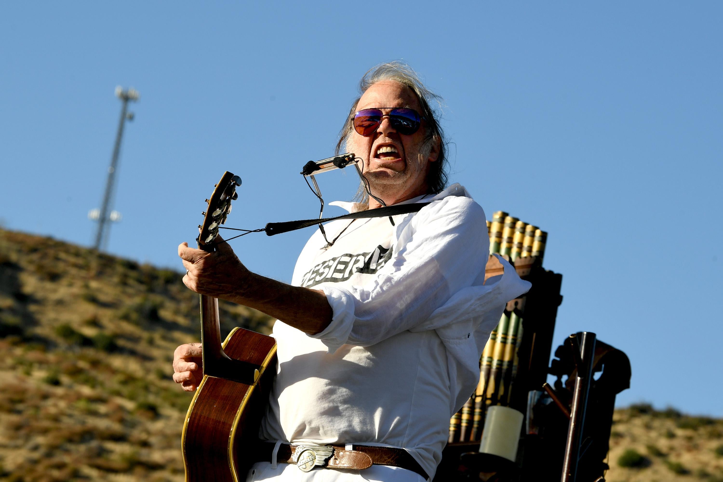 Neil Young carries a guitar and harmonica while performing onstage.