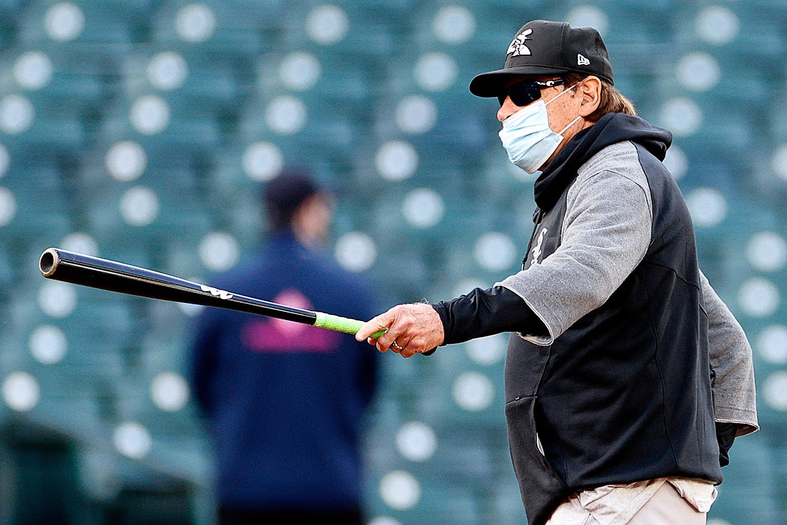 Tony La Russa holds out a bat in one hand. He is wearing a face mask that doesn't cover his nose.