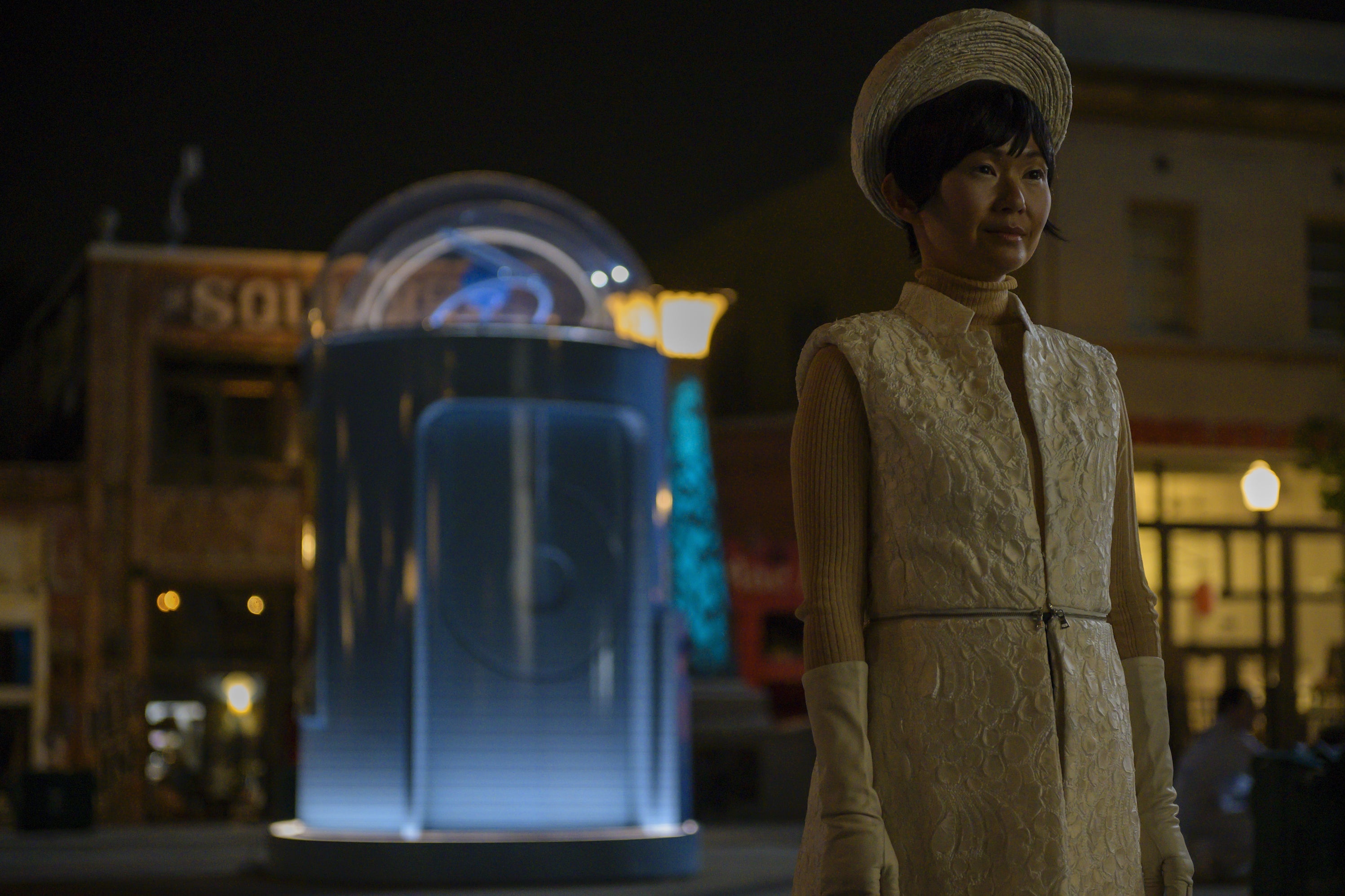 Watchmen's Lady Trieu stands in the town square of Tulsa, a glowing booth behind her.