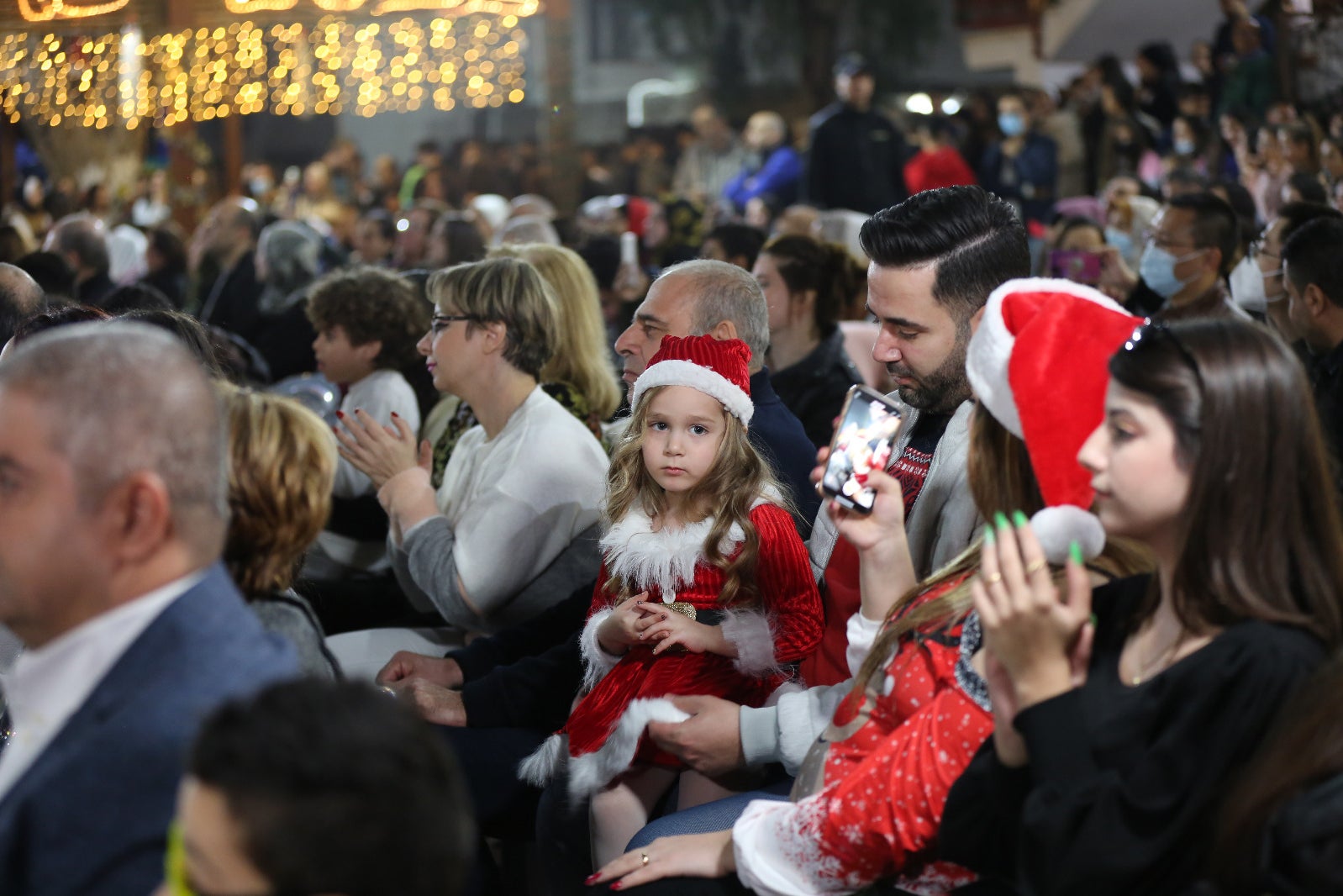 A girl in a Santa outfit and hat looks at the camera, held in her father’s lap, in the pews where everyone is dressed nicely