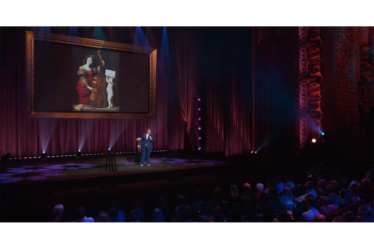 Hannah Gadsby onstage in front of a painting of St. Cecilia playing a bass viola. An angel holds her music up for her to read.
