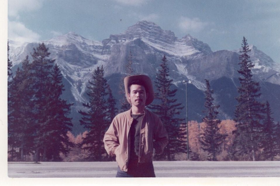 A young Japanese man wears a hat with a broad brim as he stands by the side of the road in front of a mountain range.