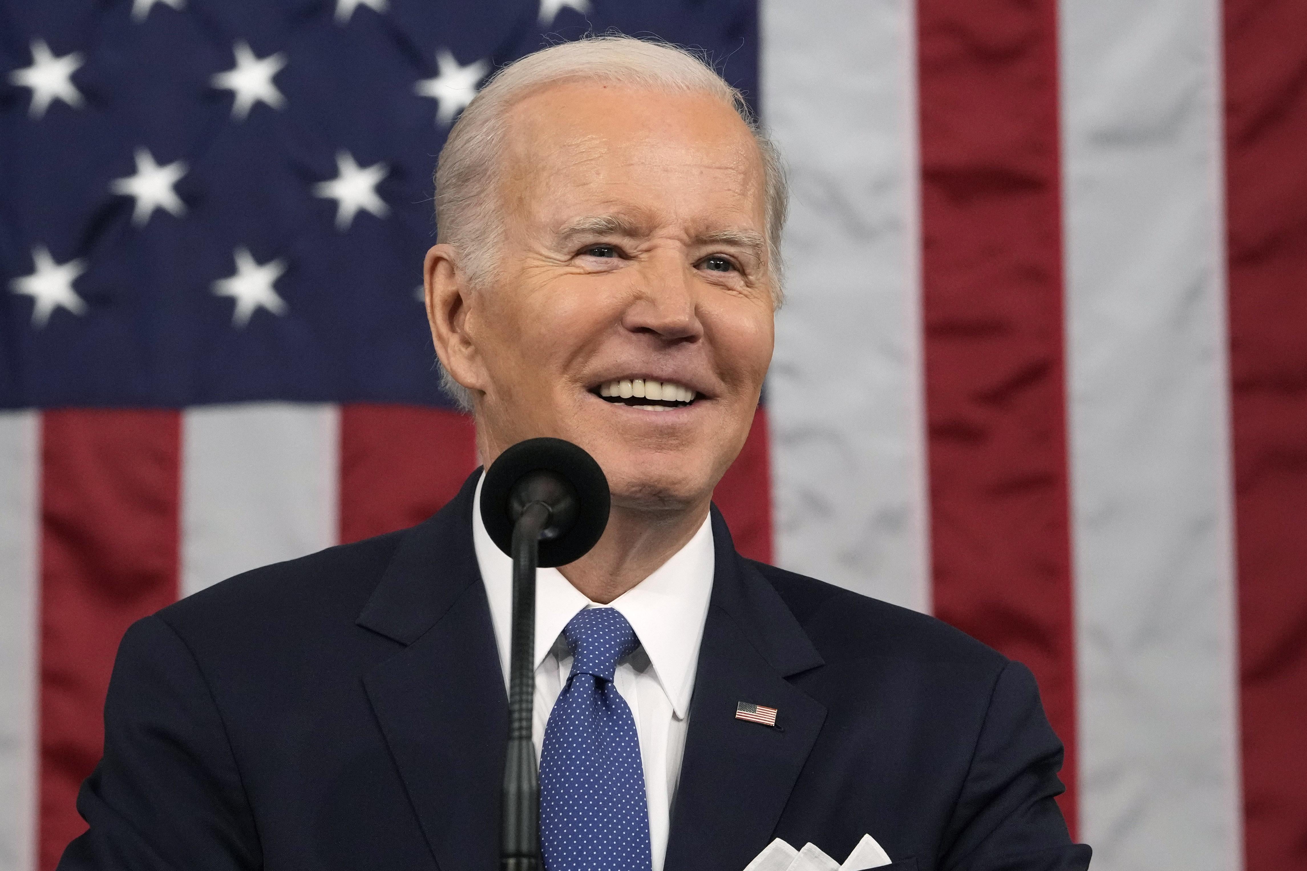 Joe Biden smiling while delivering the State of the Union, with a large American flag draped in the background