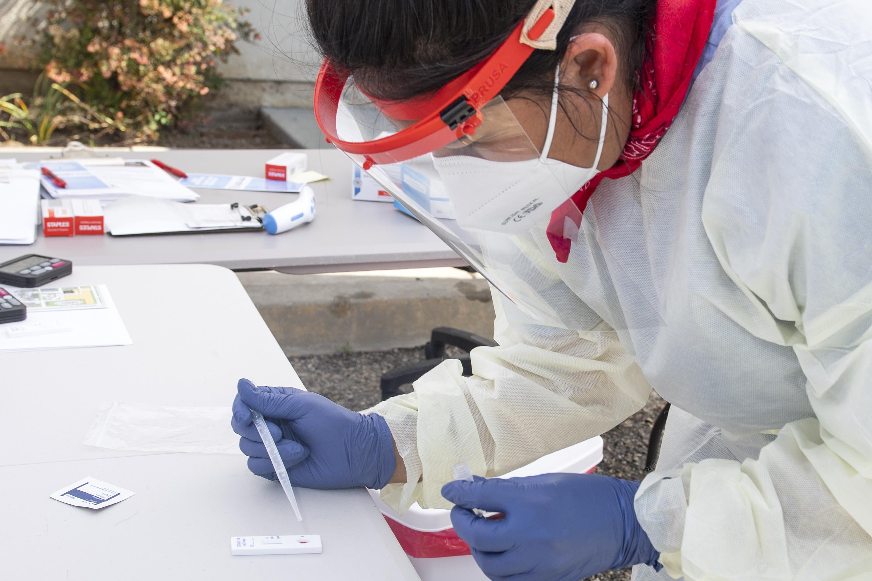A woman in protective clothing leans over an antibody test on a table under a tent.