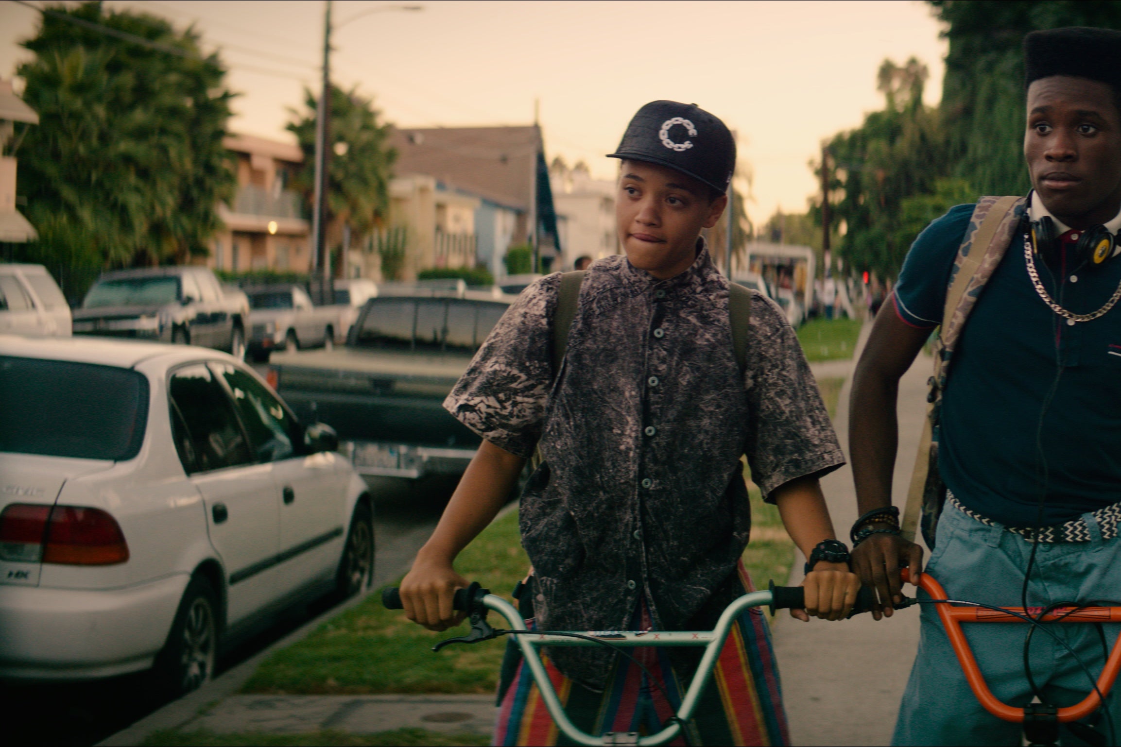 Three high-school aged kids dressed in 1990's hip-hop culture clothing, with backpacks on, pause during a neighborhood bike ride after school and stare at something off-screen. 