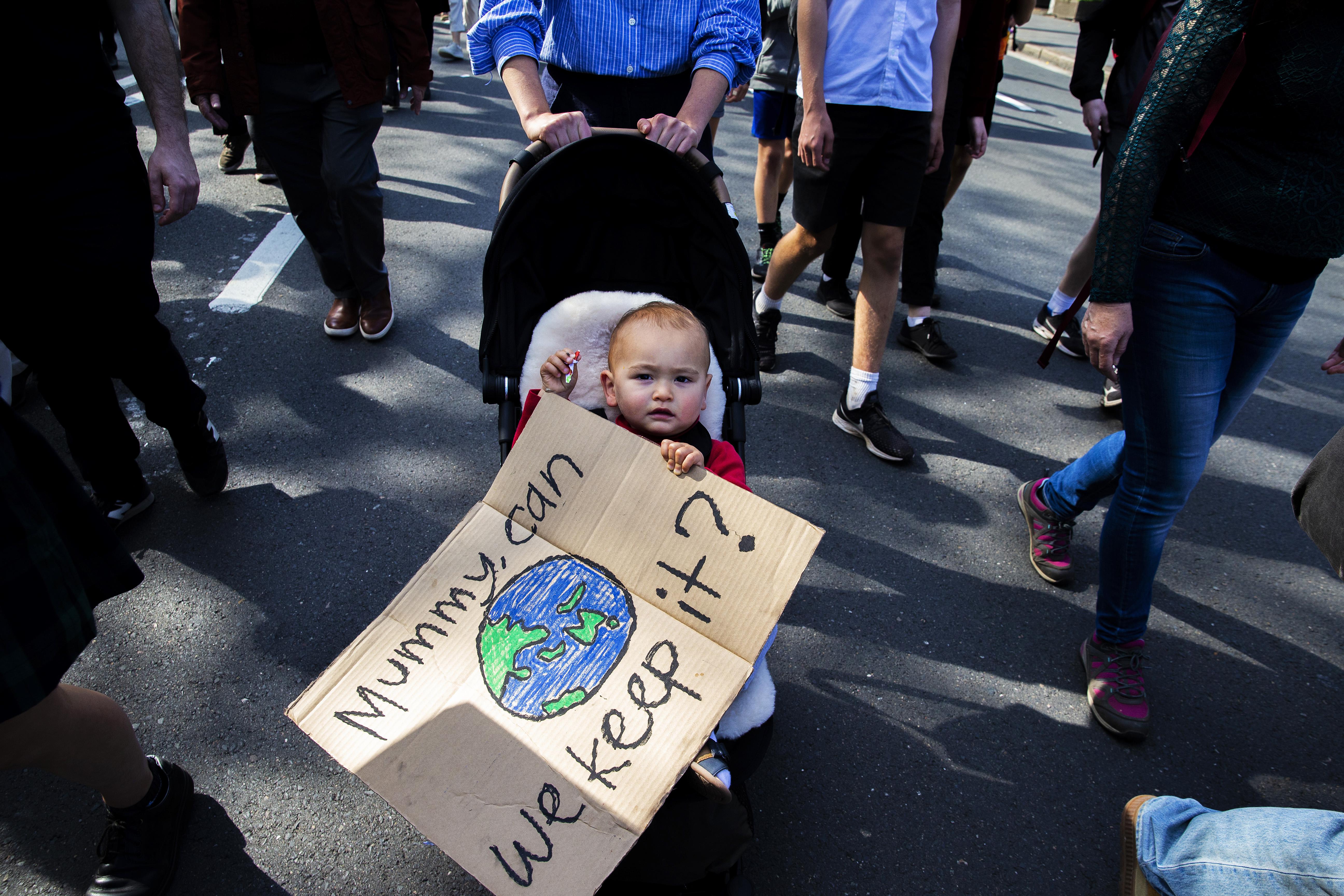 A baby in a stroller holds a sign with a drawing of Earth. The sign reads, "Mummy, can we keep it?"