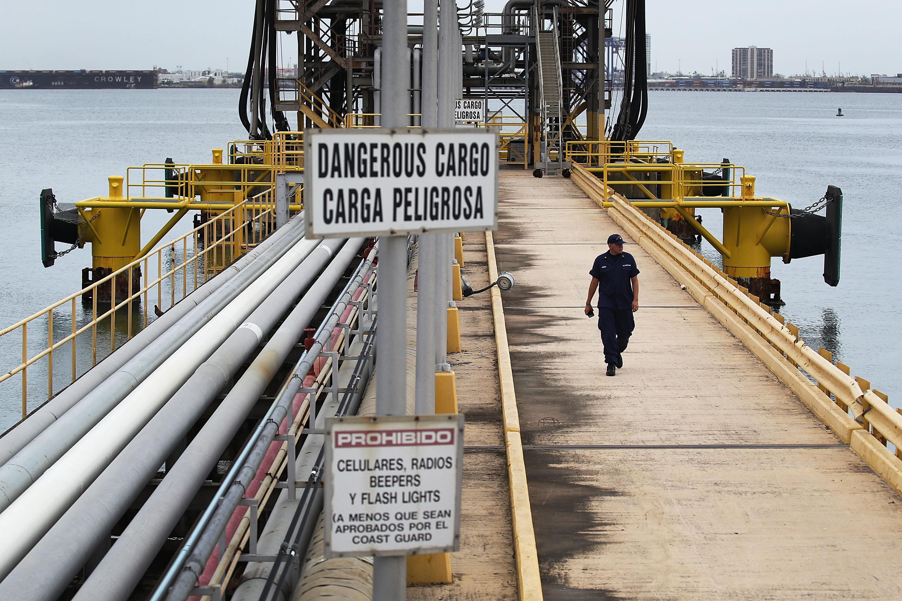 A man in a Coast Guard uniform walks down a long cement dock with oil pipes along the side. 