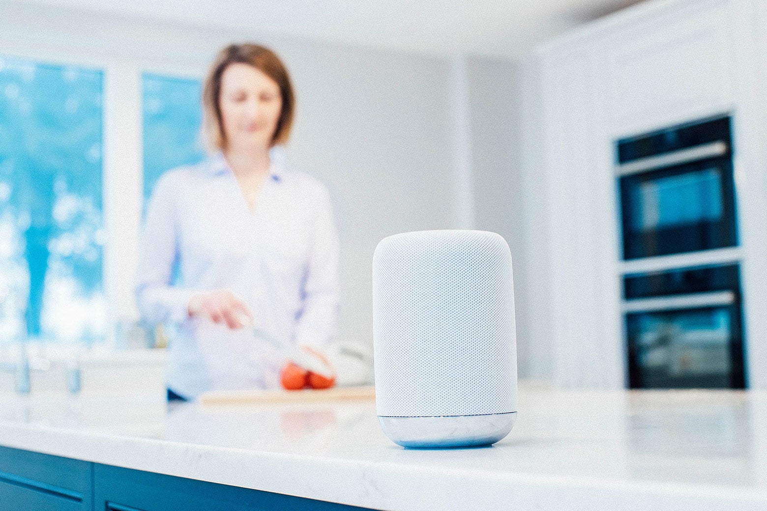 A device that looks like an Apple HomePod rests on a kitchen counter while a woman prepares food in the background.