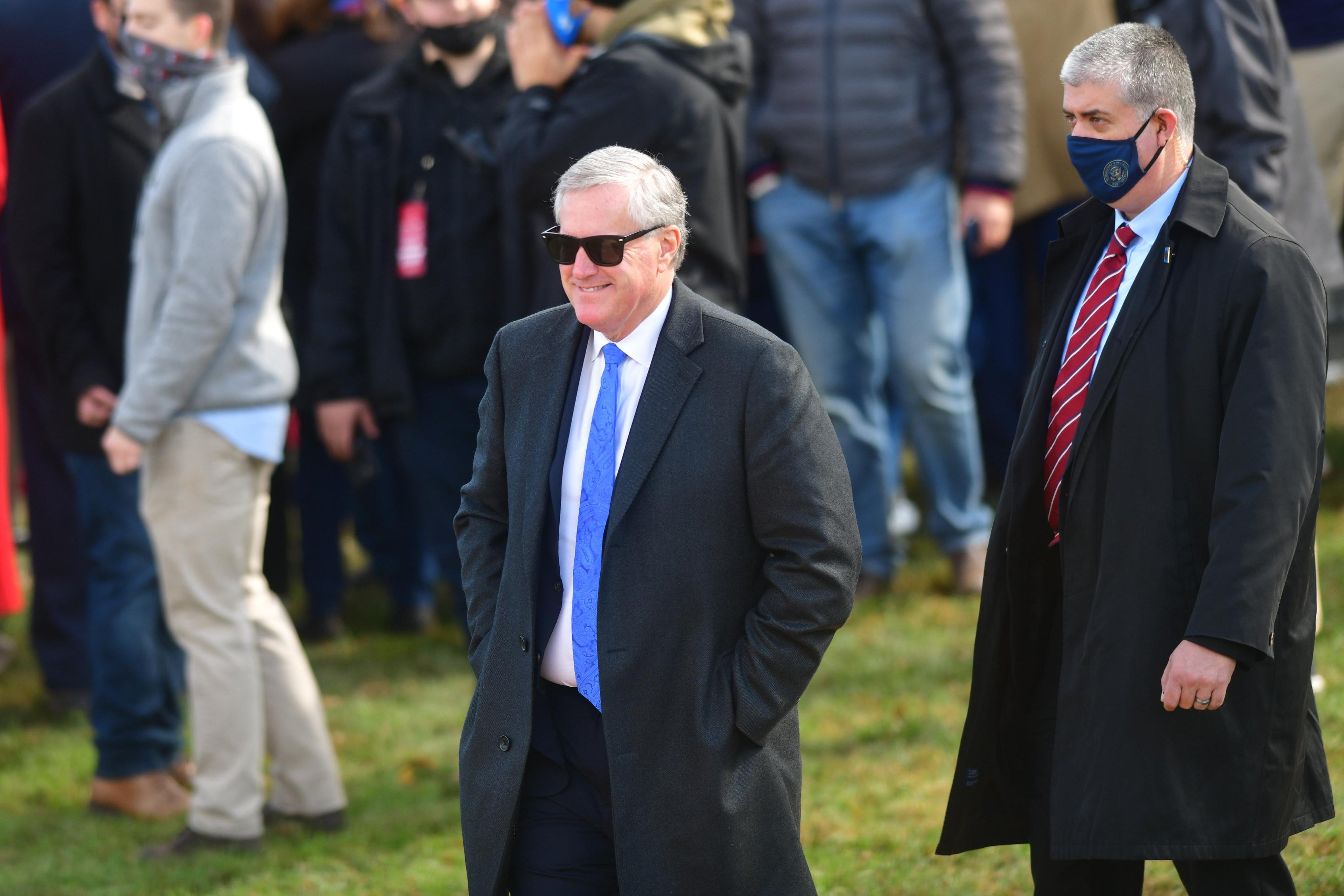Mark Meadows smiles as he walks with his hands in the pockets of his trenchcoat in front of a crowd of Trump supporters standing on grass