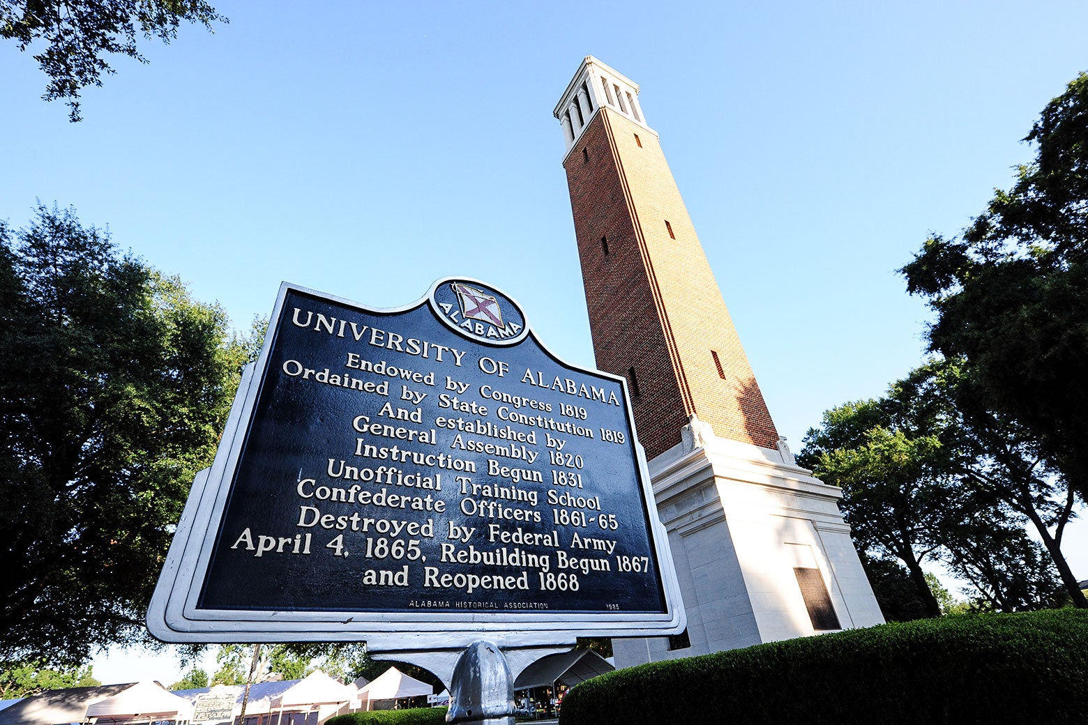 A sign introducing the University of Alabama is seen in front of a tall bell tower.