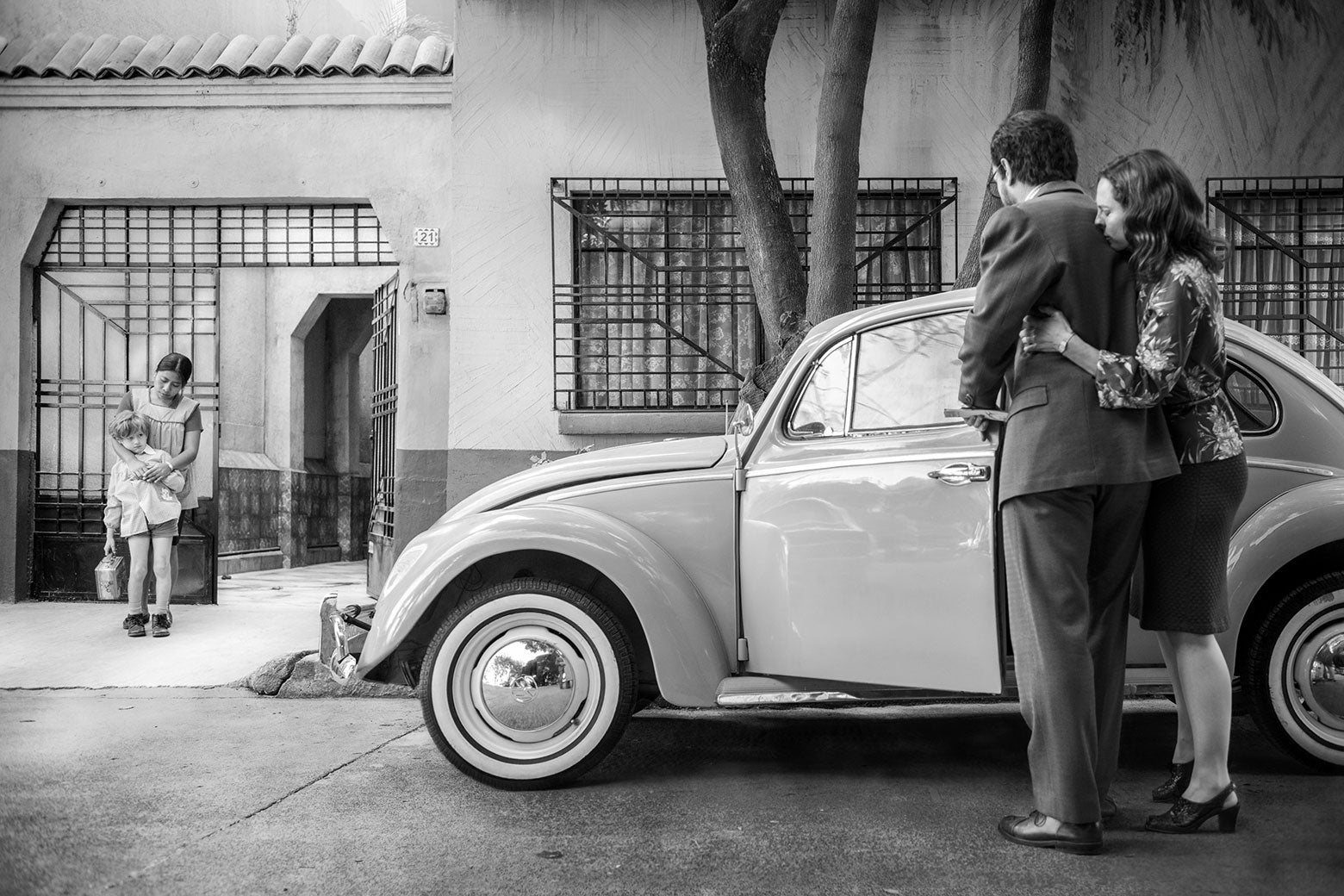 In a still from Roma, a woman clings to her husband beside their car, and a maid comforts their child outside the gate of their home.