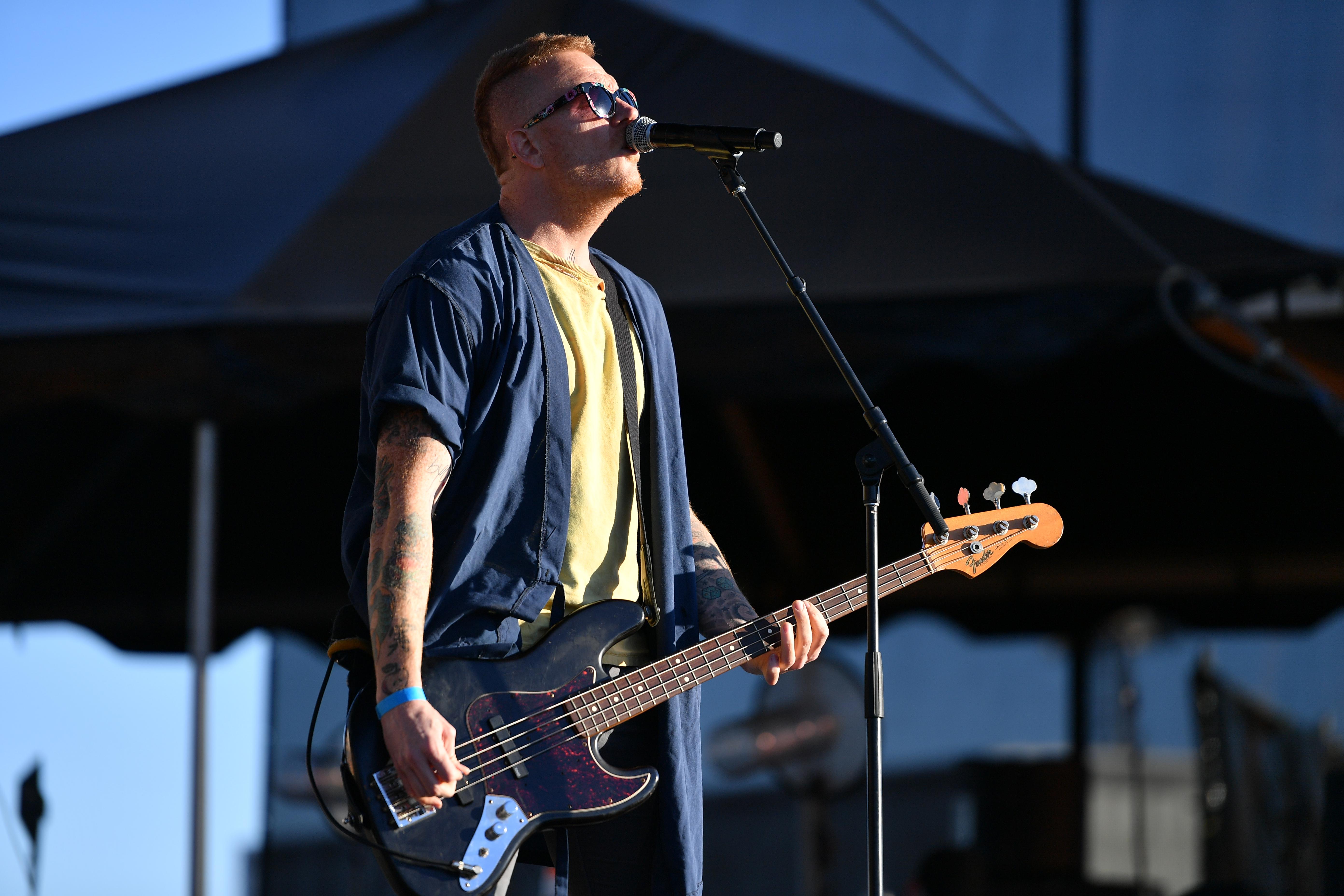 A man wearing a blue button-down shirt over a yellow T-shirt stands onstage playing a bass and singing into a microphone.