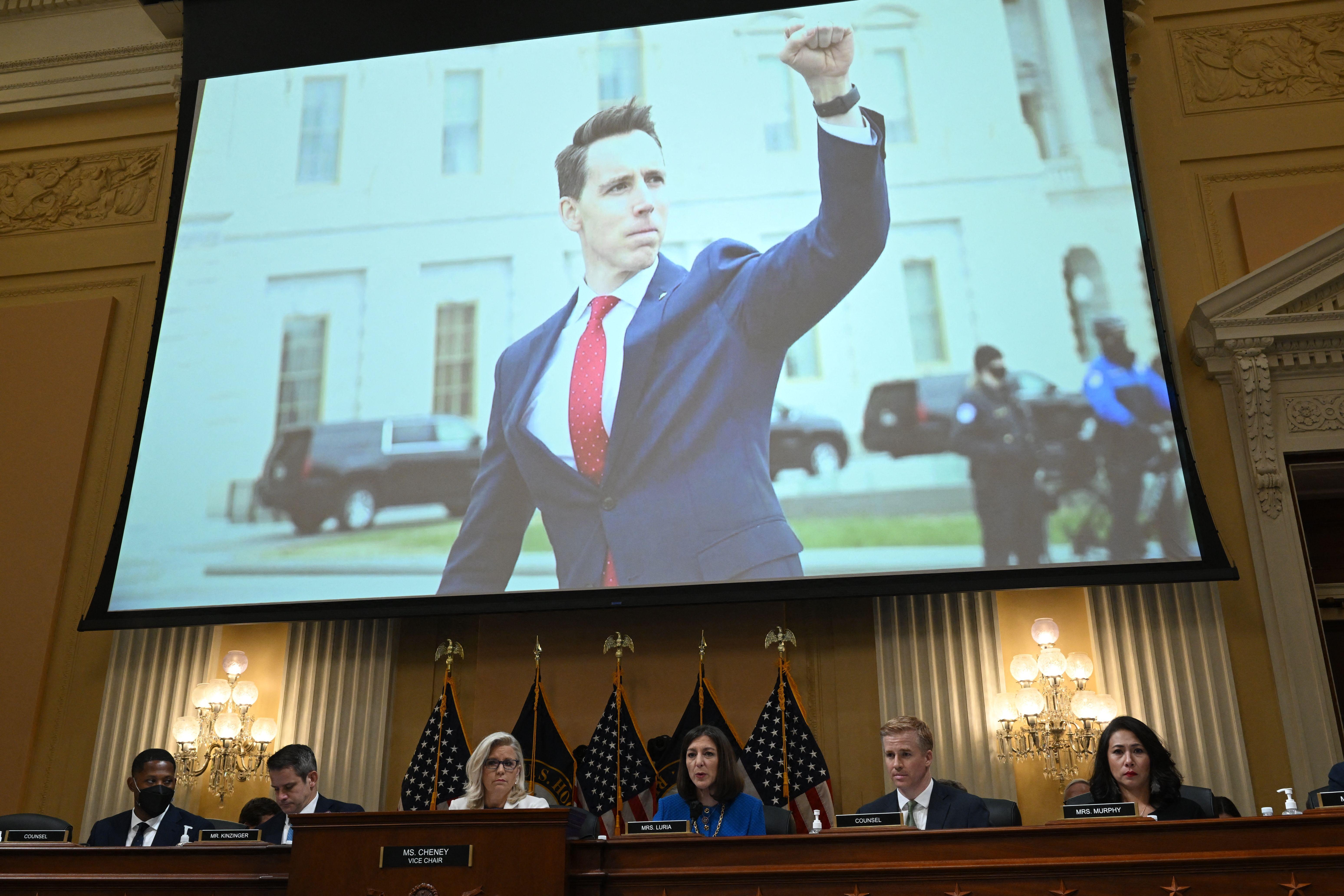 Hawley raising his fist in a blue suit and red tie is projected to a hearing room.