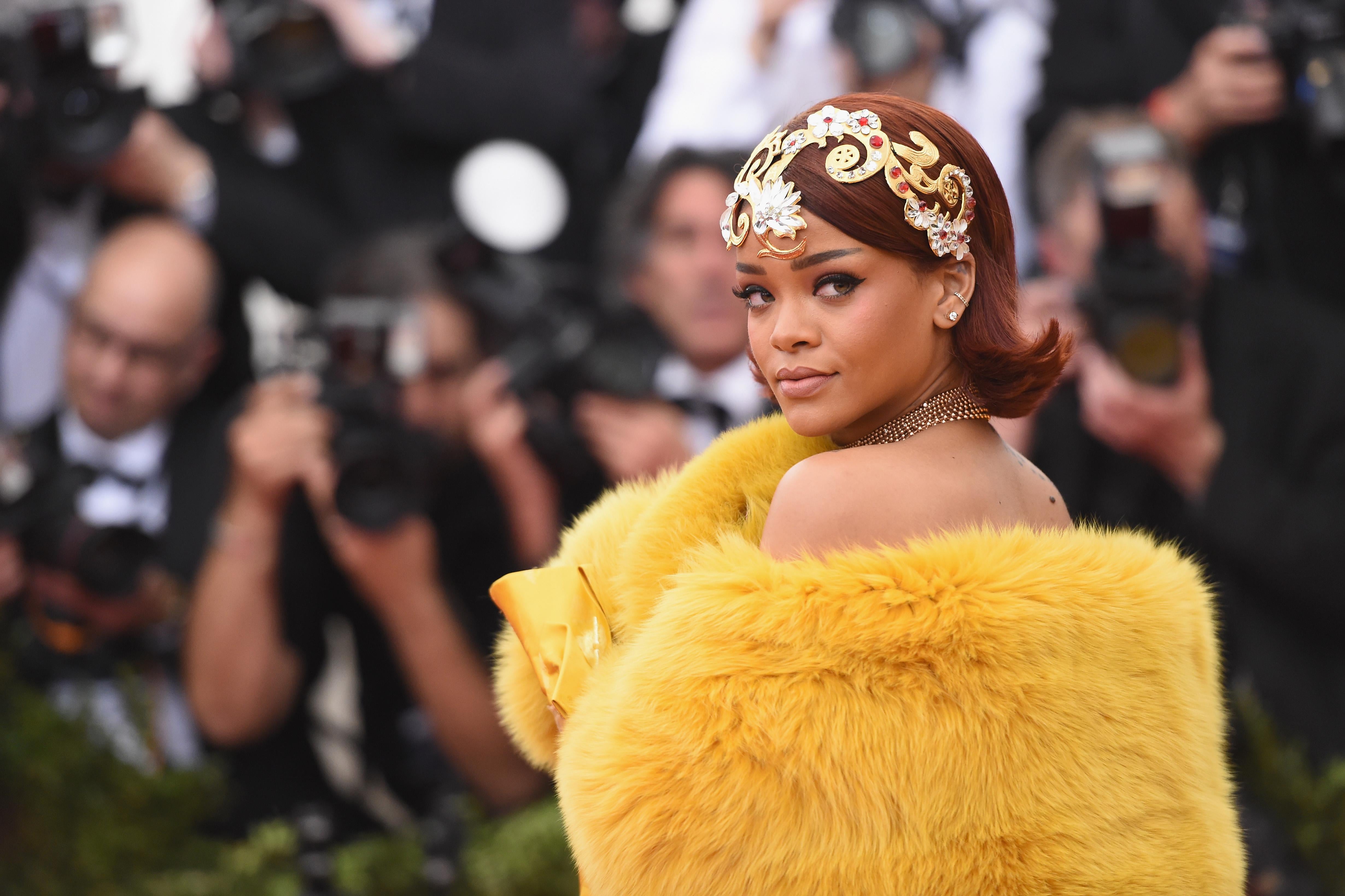 Rihanna looks over her shoulder. She wears a dress that, at the top, resembles a yellow fur capelet.