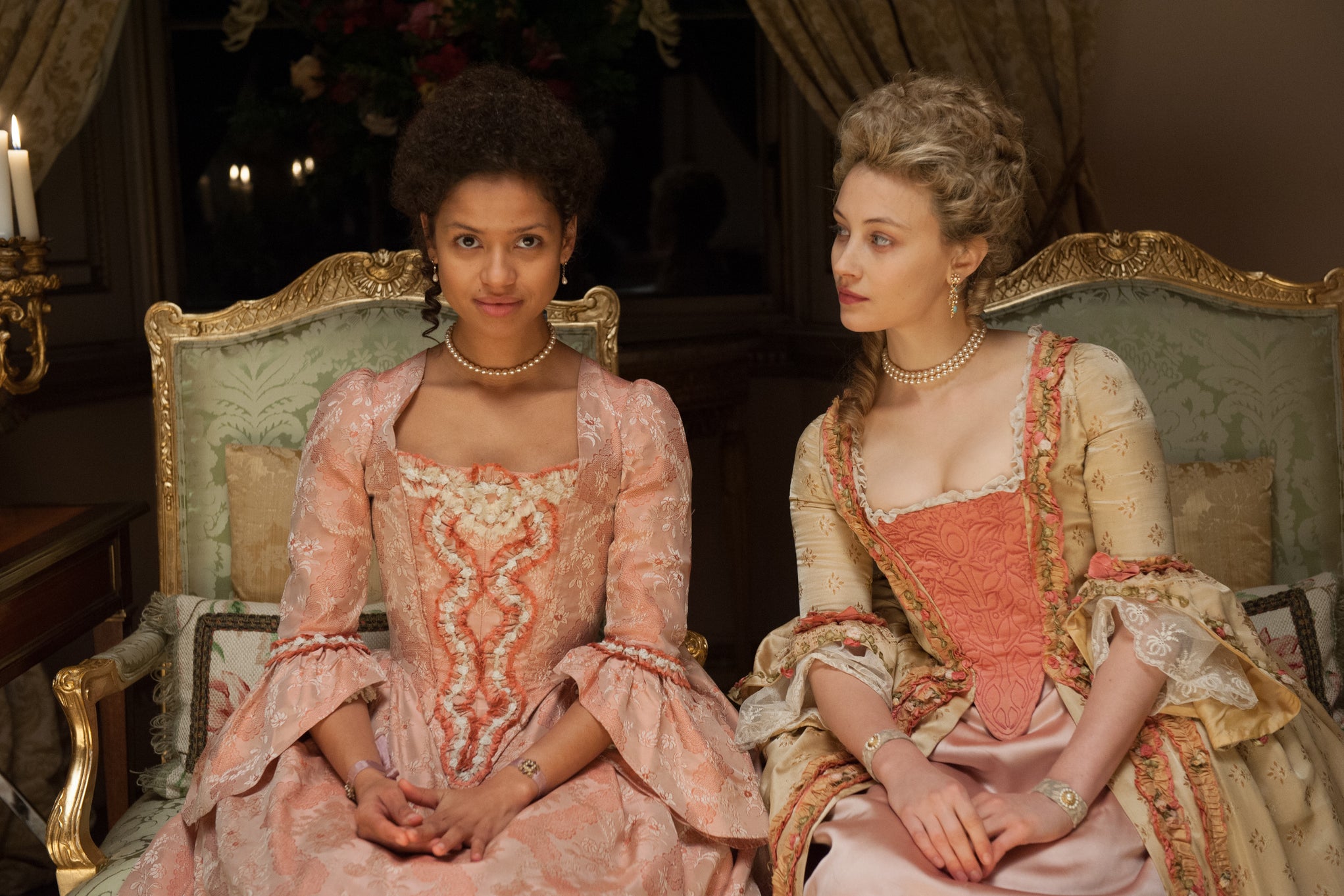 In 18th century England, a young Black woman dressed in Georgian Era garb, looks ahead with a slight smile on her face, as she sits in an ornate drawing room next to a young white woman, also dressed in Georgian Era garb, who is looking at her with curiosity. 