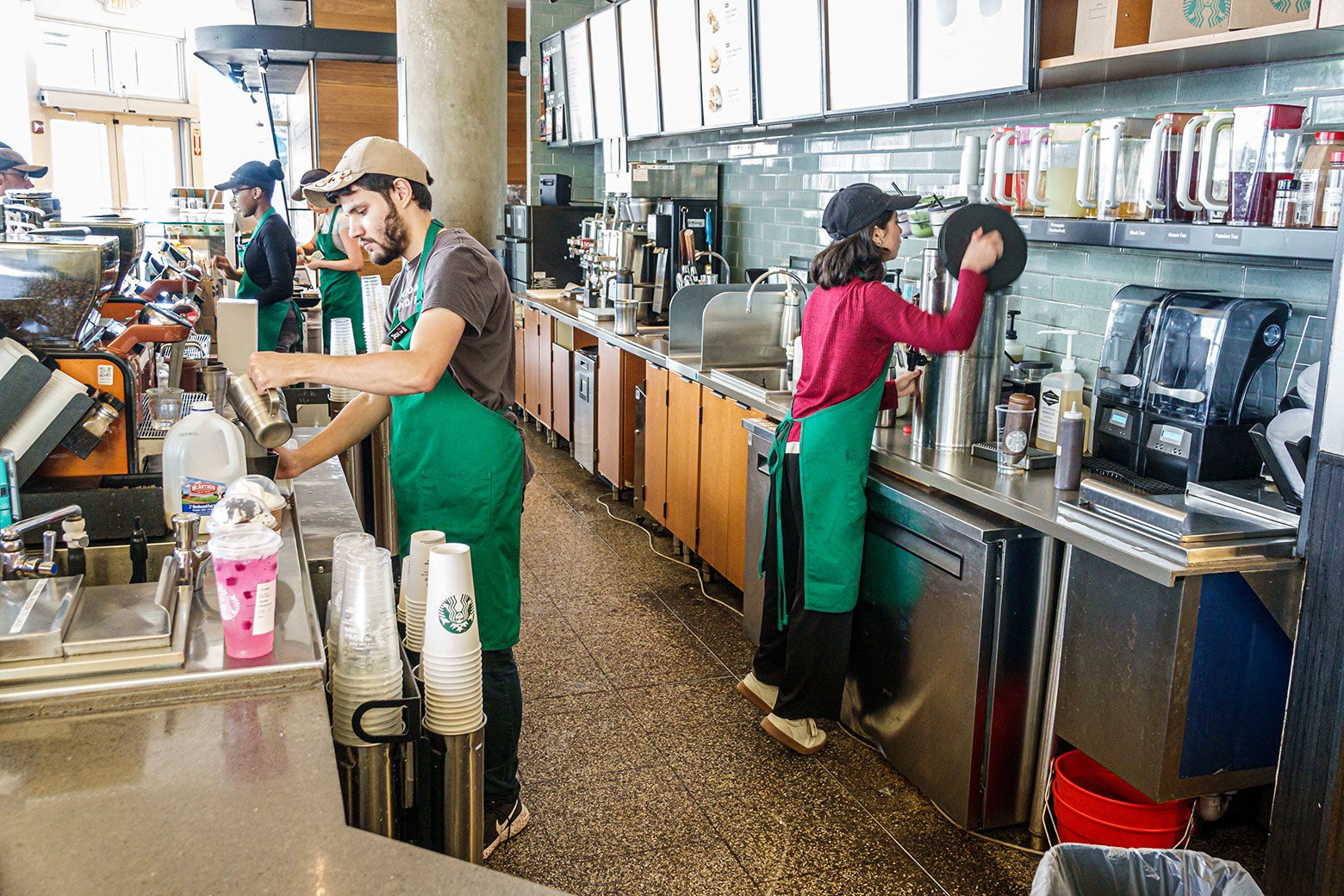 Behind the counter of a Starbucks one barista is making a huge pink drink and another has to stand on tiptoes to see into the machine she is using. 