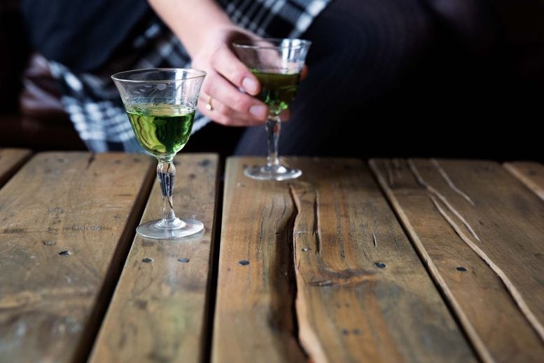 Two glasses of green Chartreuse on a table, with a hand reaching toward one glass
