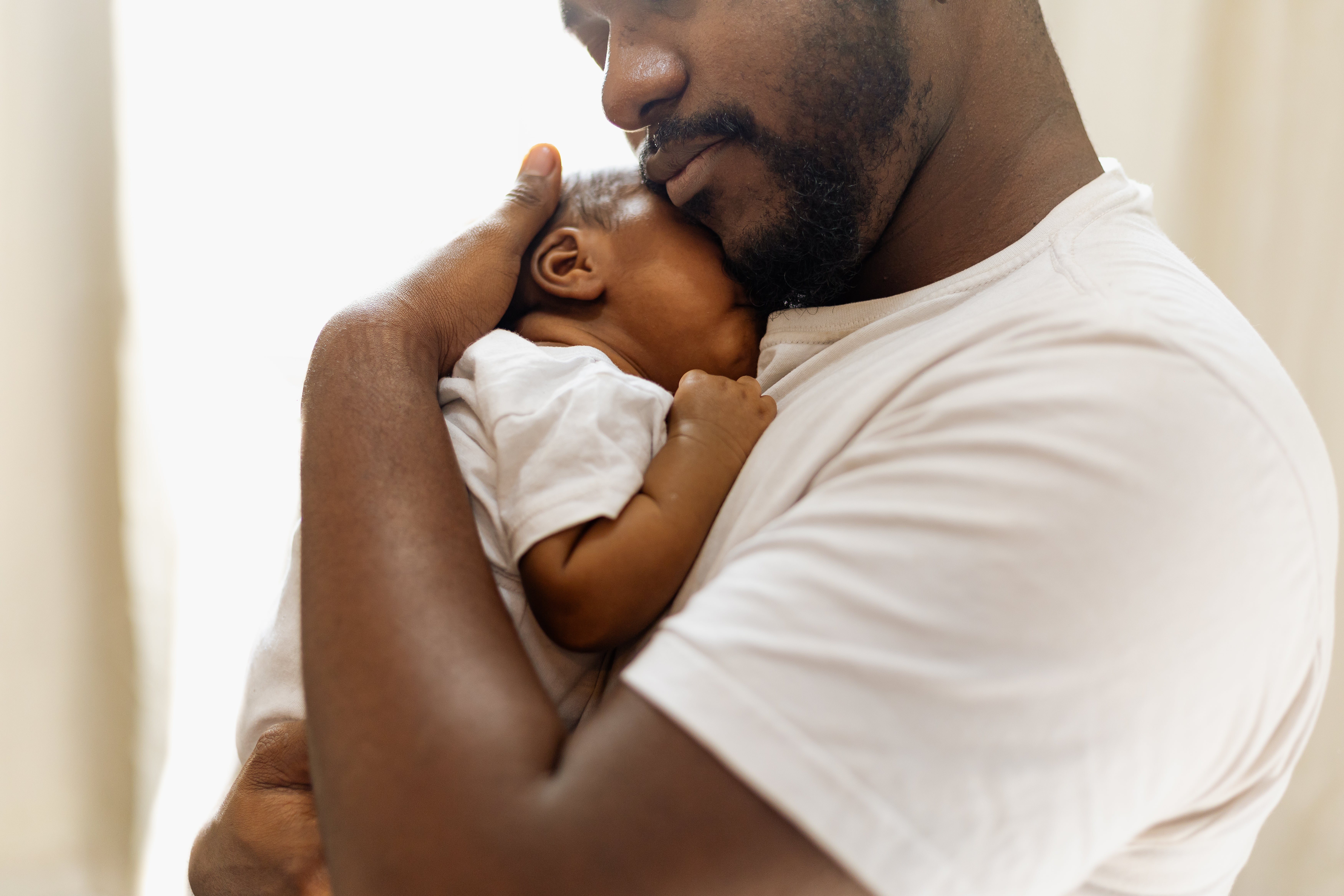 A bearded man is cradling his infant to his chest and leaning his chin on the baby's head.