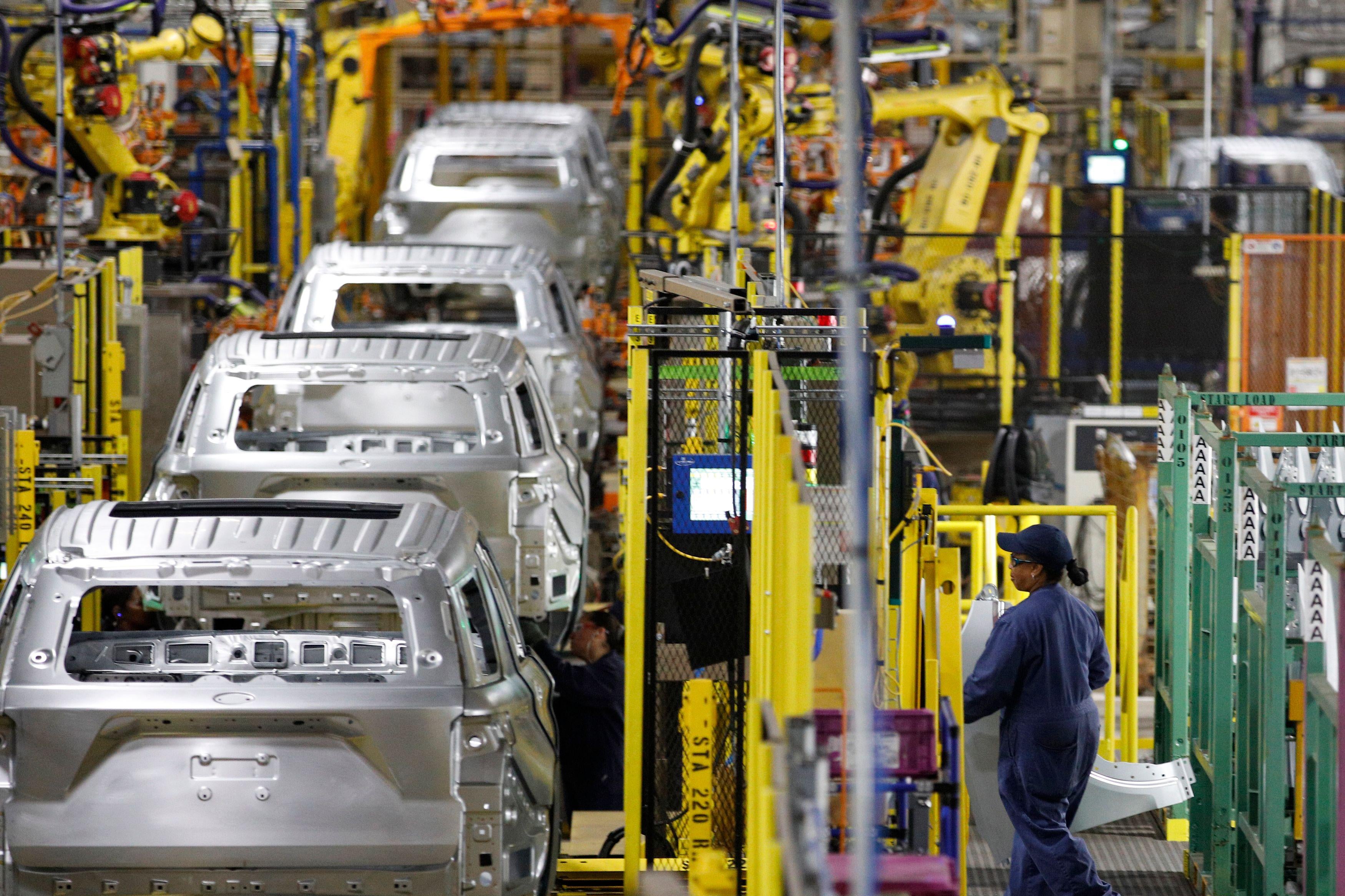 Unfinished cars are lined up on an assembly line on the left. Machinery surrounds them. A woman in a blue uniform walks toward them on the right.