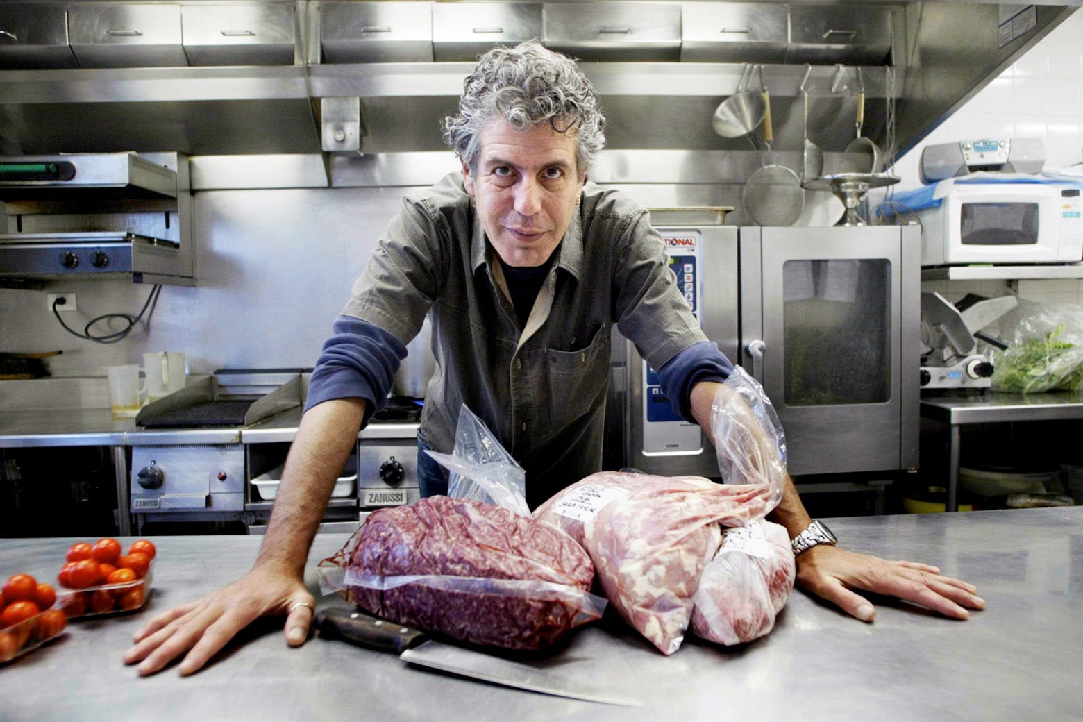 Anthony Bourdain leans over a counter in a stainless-steel kitchen. There are bags of meat and a knife on the counter.