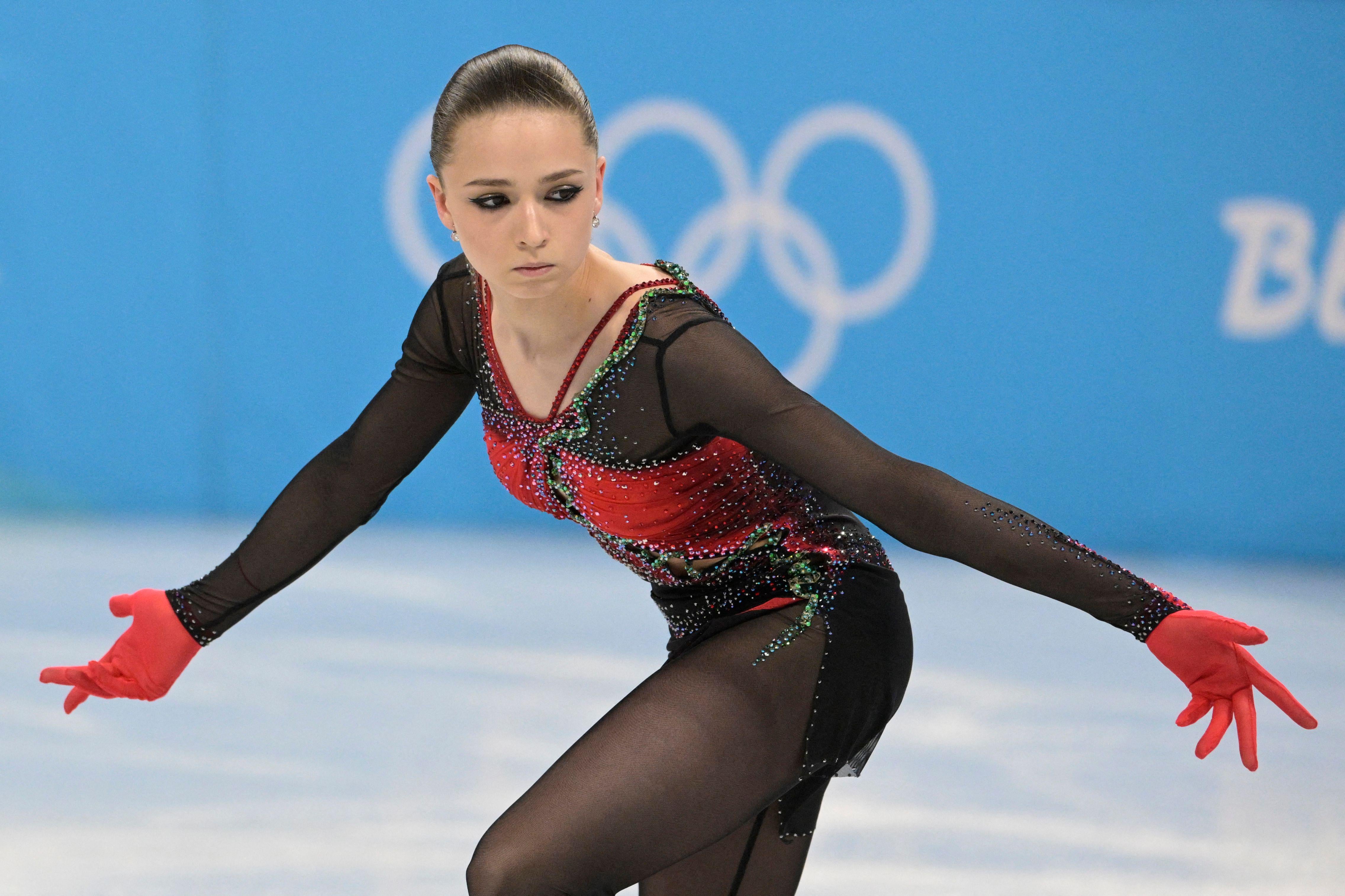 Valieva turning on the ice, stony-faced with her arms extended outward and gloved palms up